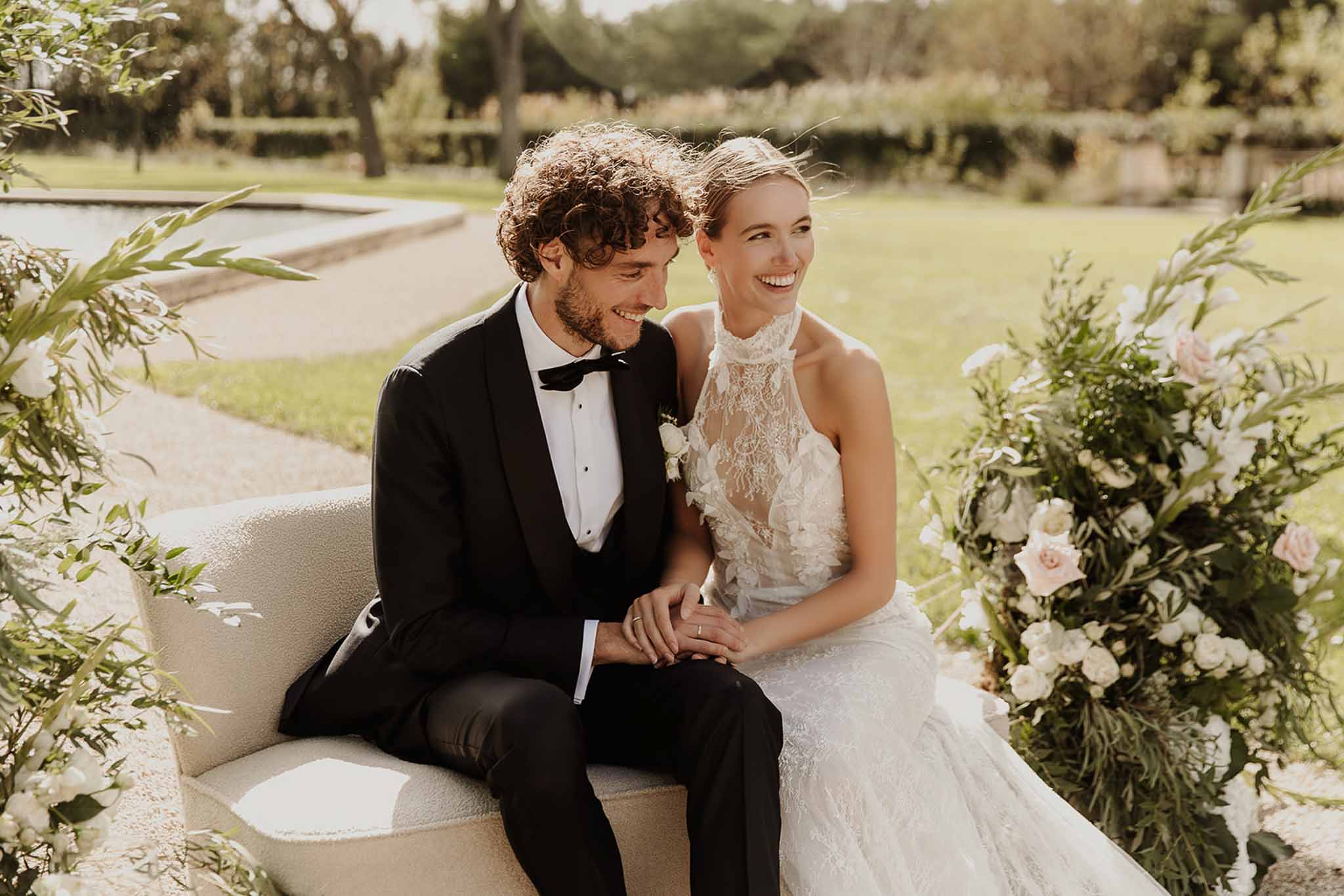 Couple laughing on cream sofa flanked by blush and white rose arrangements in garden with reflecting pool