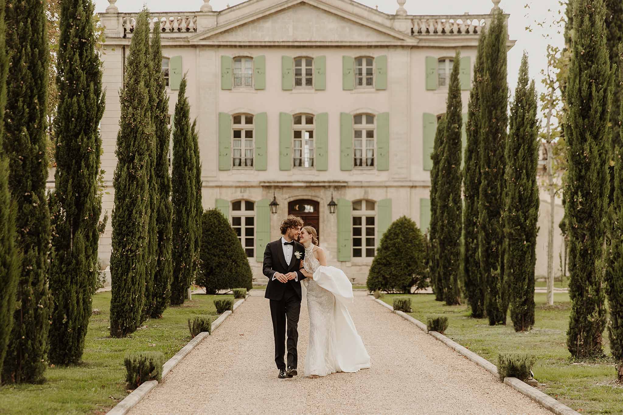 Couple walking toward camera on cypress-lined driveway bride in lace gown with detachable train pink stone chateau behind