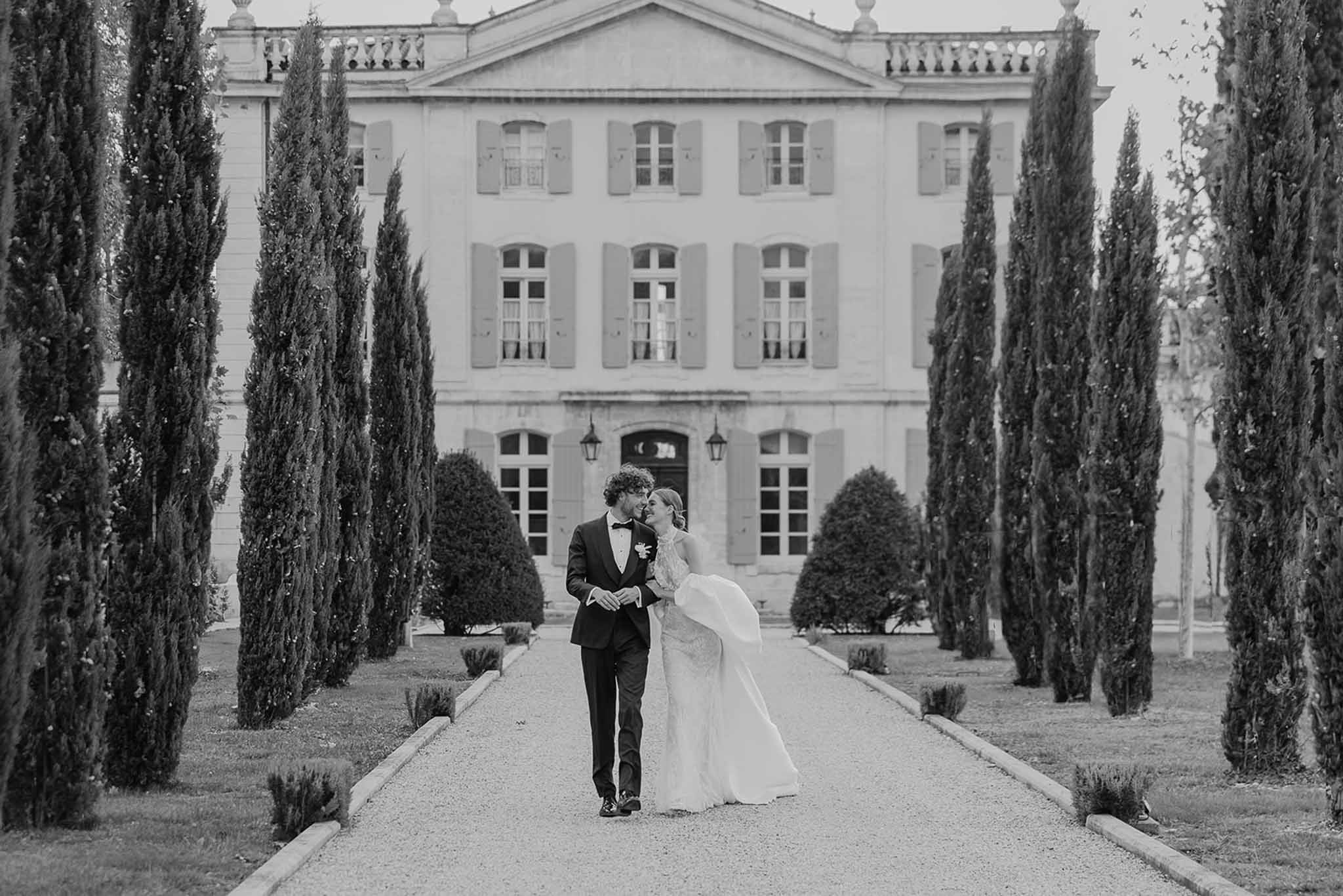 Black-and-white portrait of bride and groom walking along cypress-lined gravel allee toward French chateau