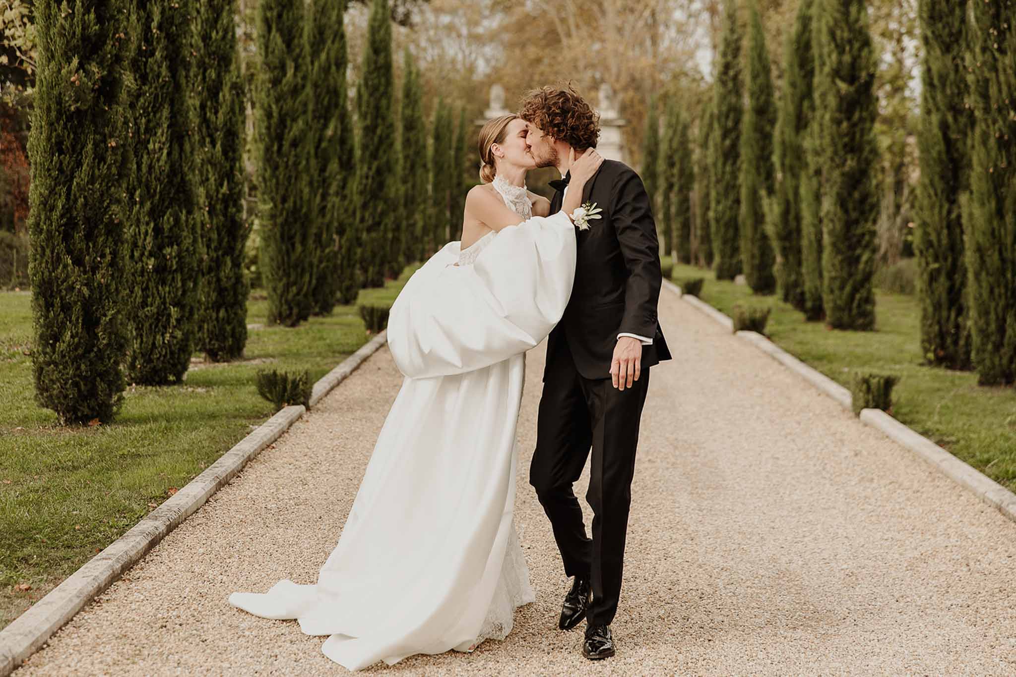 Couple kissing on cypress-lined gravel allee, bride in white lace halter gown with balloon sleeves and full train
