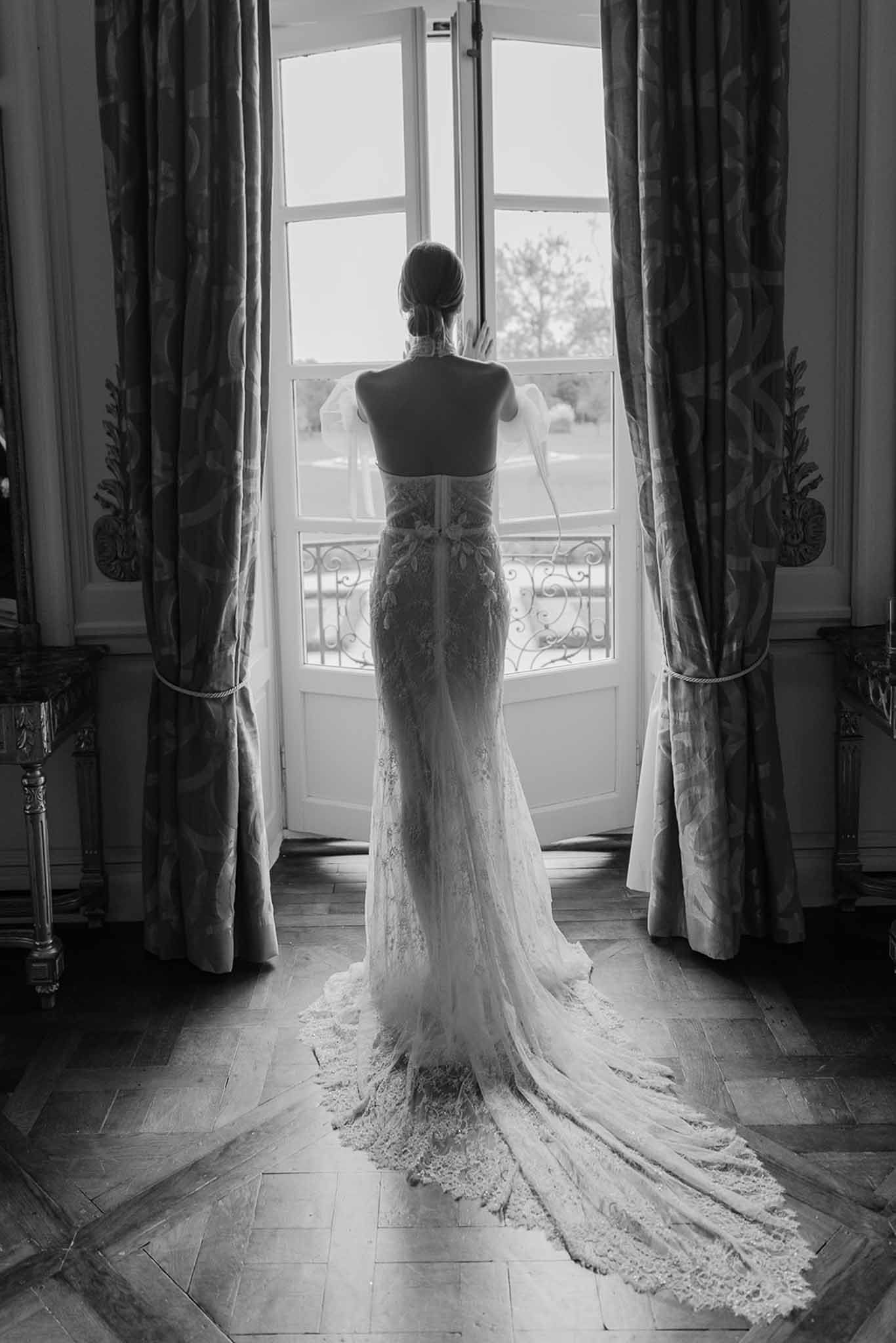 Black and white portrait of bride from behind showing lace open-back gown and cathedral train in chateau doorway