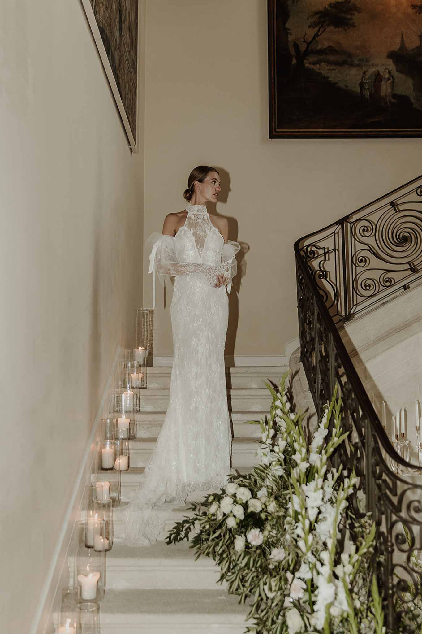 Bride in white lace halter gown on marble staircase lined with candles and white rose arrangements