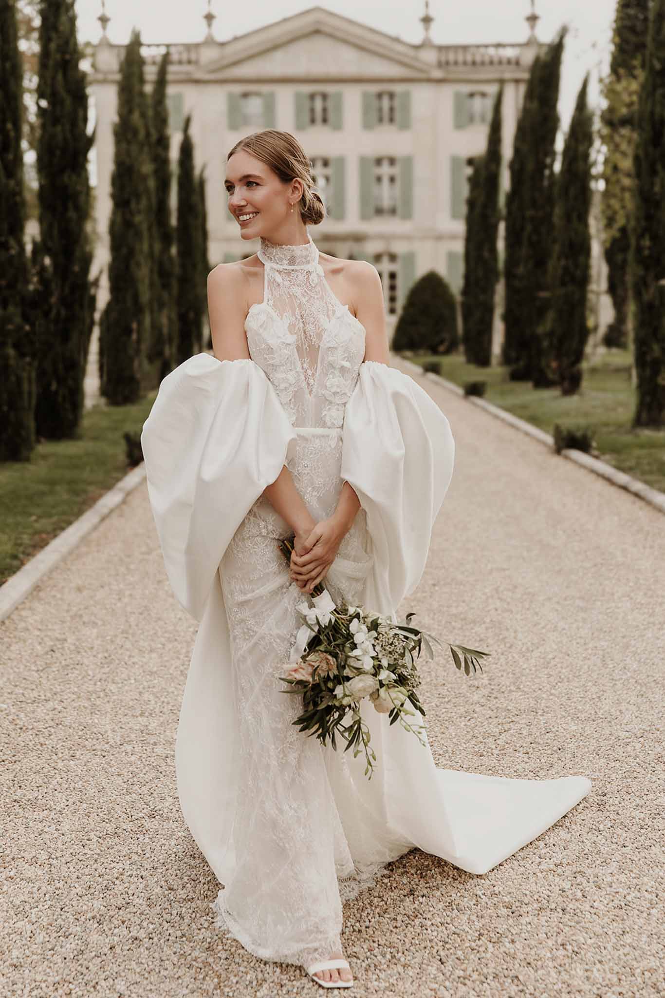Bride in ivory lace halter gown with white structured cape holding bouquet on chateau gravel driveway