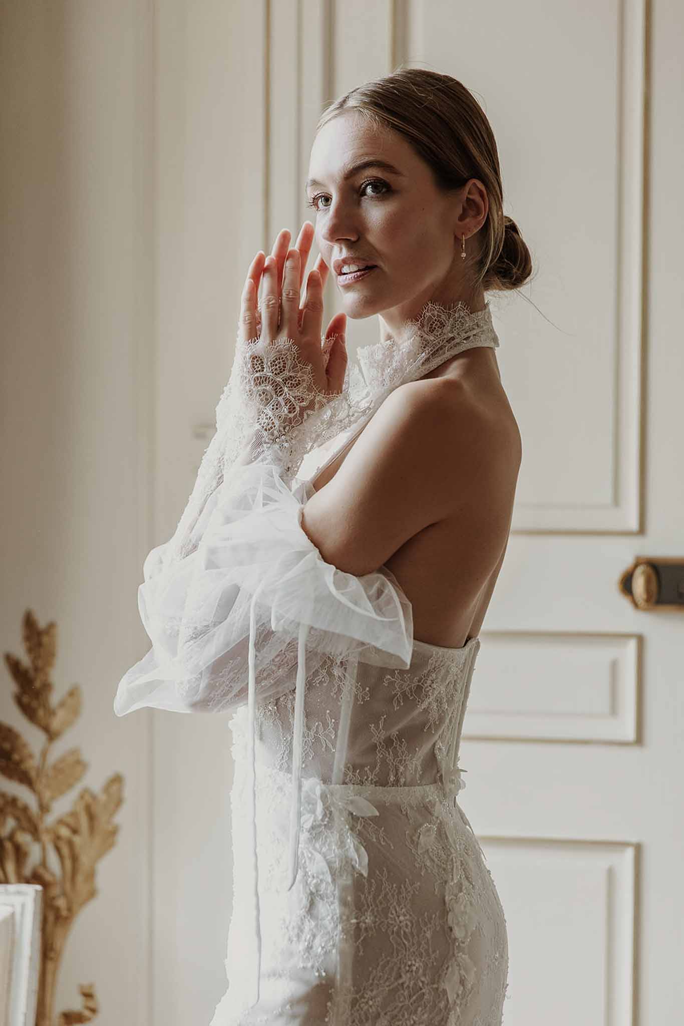 Bride in ivory halter lace gown with tulle bow sleeves posed against cream panelled walls with gold trim