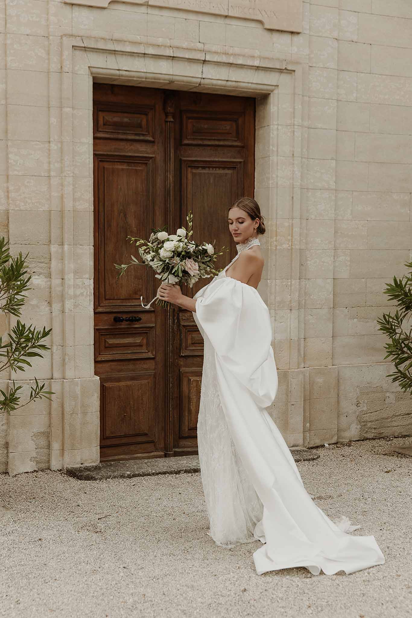 Bride in profile with draped satin overskirt train holding garden rose and peony bouquet before limestone chateau doors