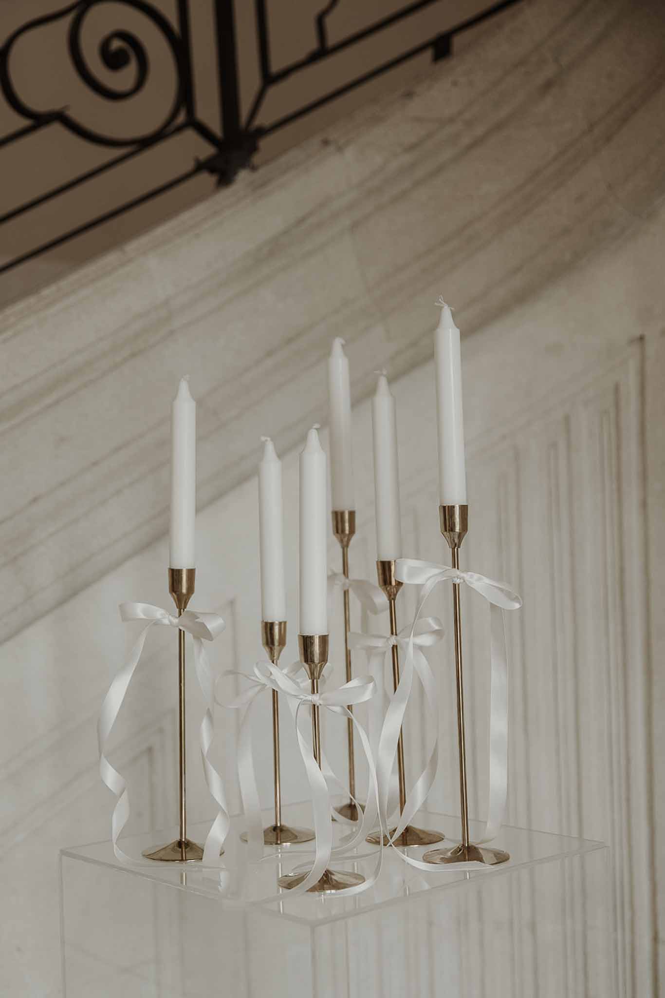 Six white taper candles in brass candlesticks tied with satin ribbon bows on acrylic plinth