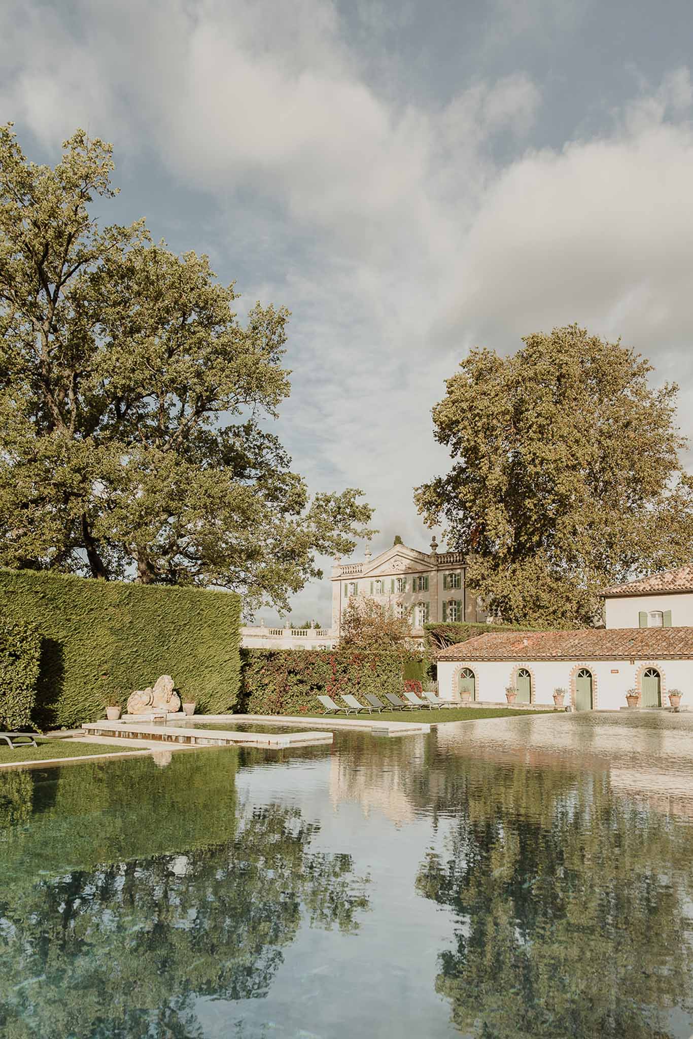 French chateau estate with rectangular reflecting pool, sun loungers, clipped hedges, and stone lion sculptures