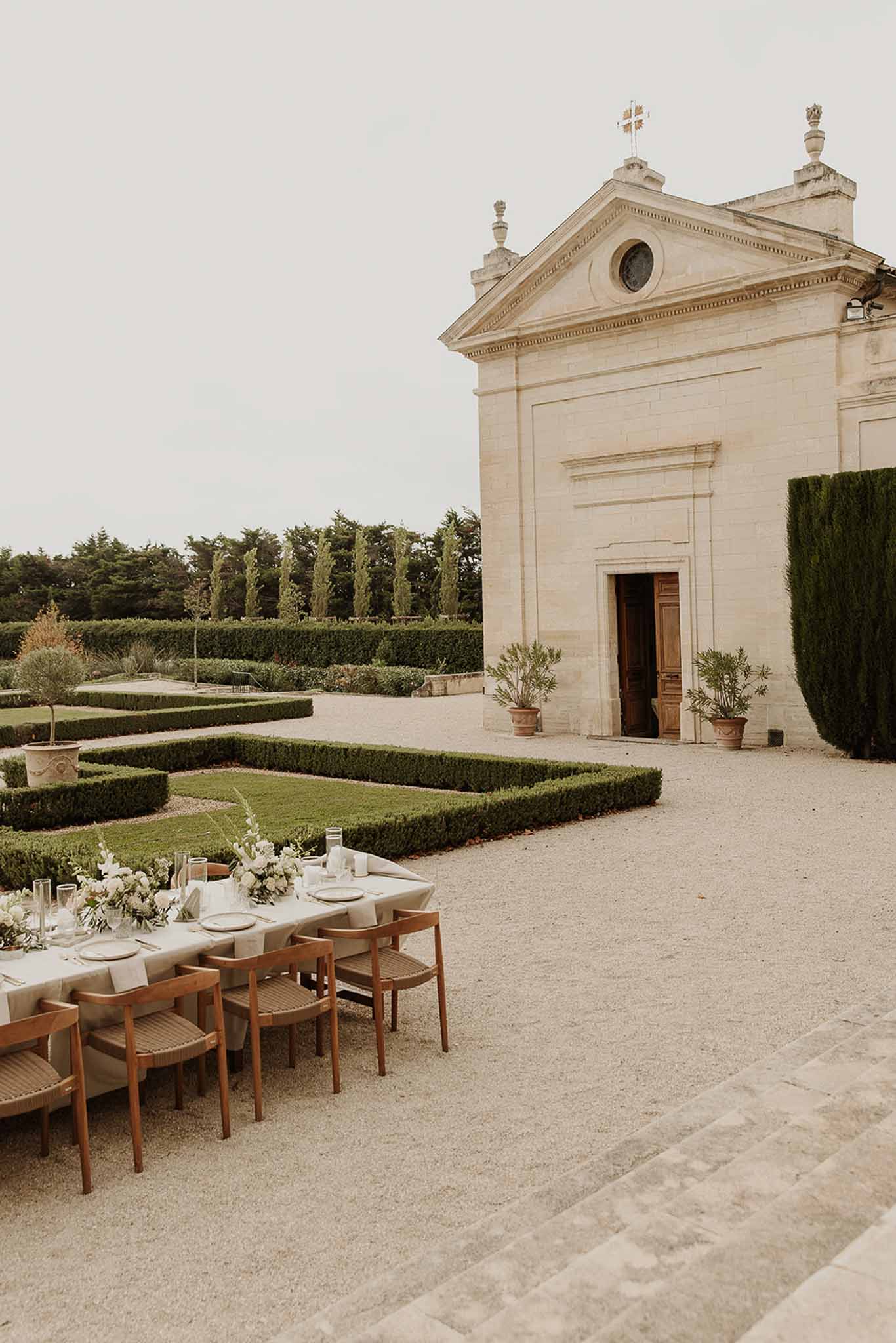Long table with cream florals and wooden chairs before limestone chapel with boxwood parterre garden
