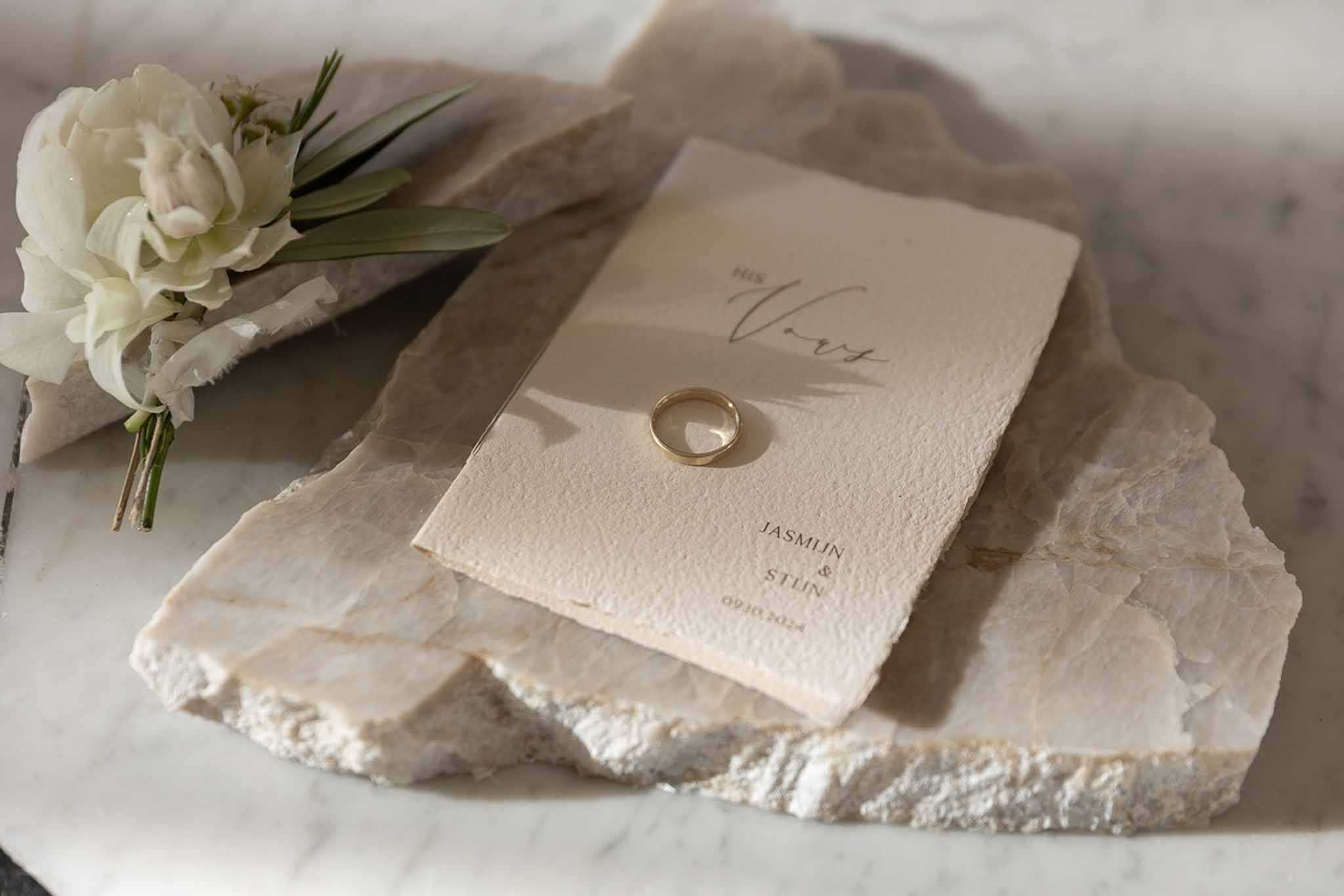 Flat lay of deckle-edged vow booklet gold wedding band and ranunculus boutonniere on stone slab