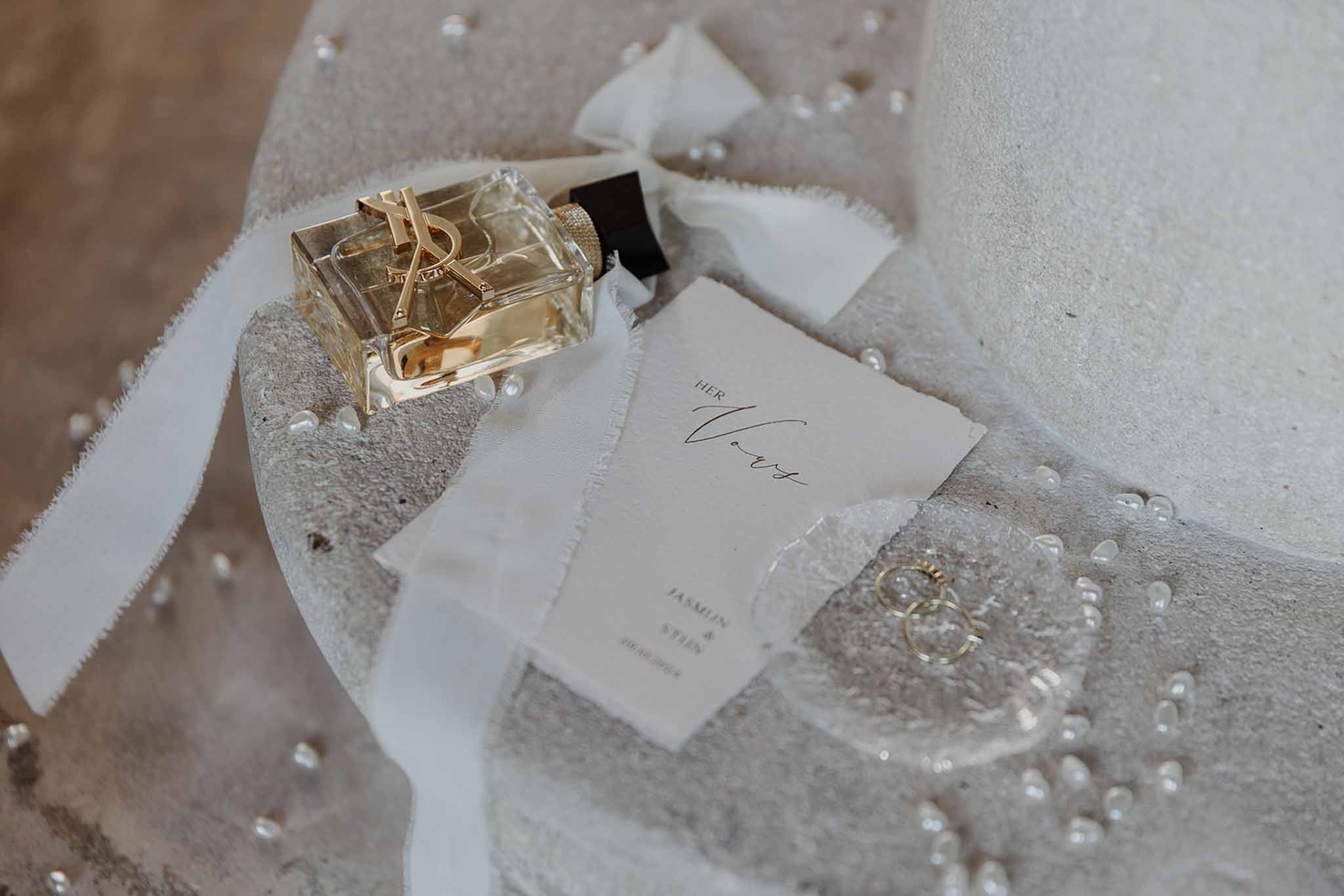 Flat lay of YSL Libre perfume, gold wedding bands, silk ribbon, and personalised vow card on stone surface with loose pearls