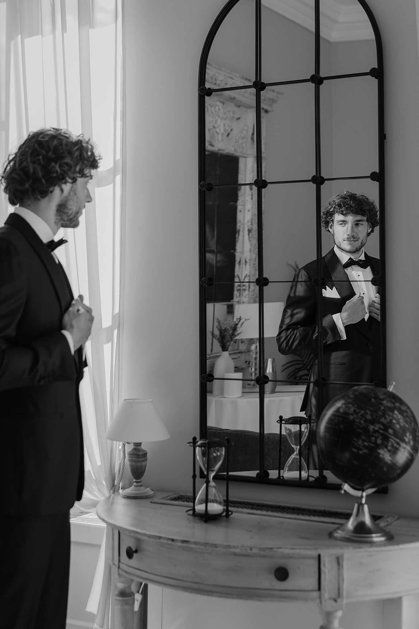 Groom adjusting bow tie in front of arched iron-framed mirror in black and white