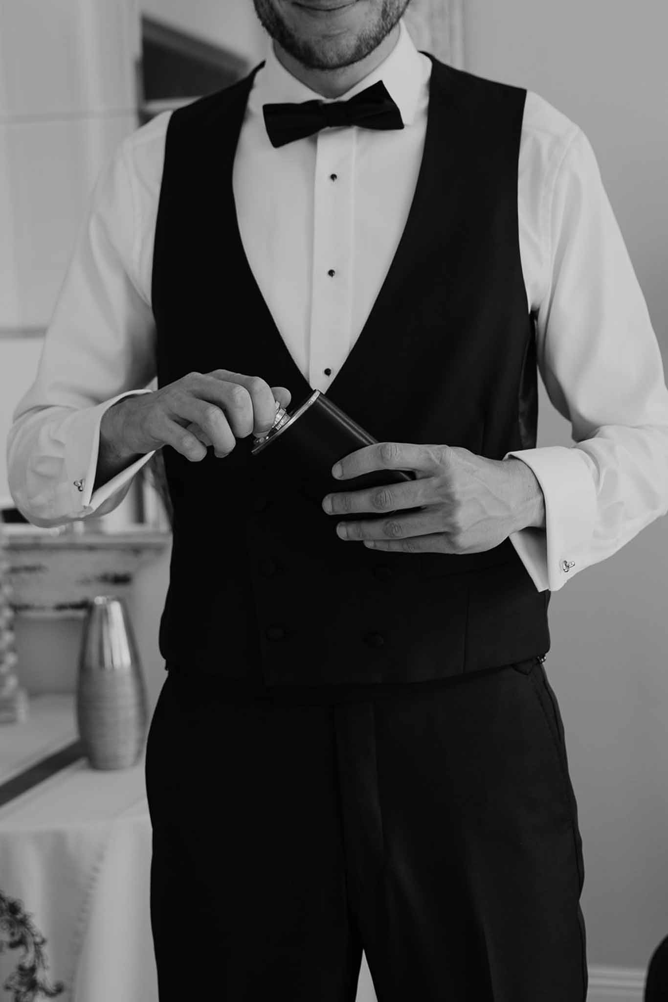 Black and white torso shot of groom in tuxedo waistcoat and bow tie opening silver hip flask