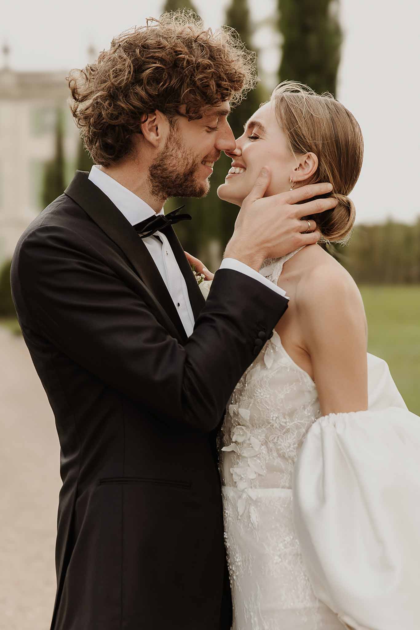 Close-up groom cupping brides face both smiling bride in floral applique gown with structured bow at chateau grounds