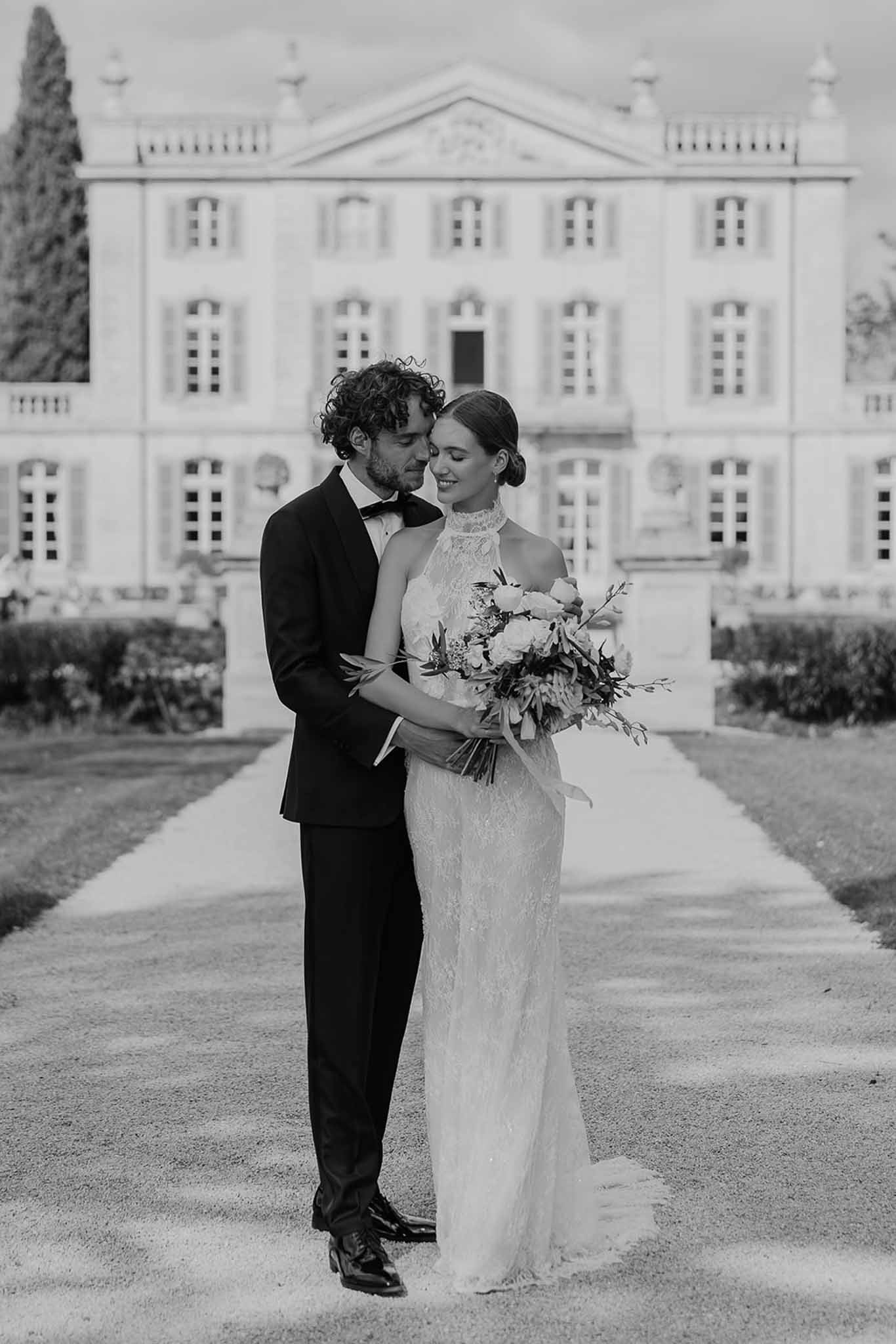 Black-and-white portrait of groom embracing bride with bouquet in front of neoclassical chateau facade