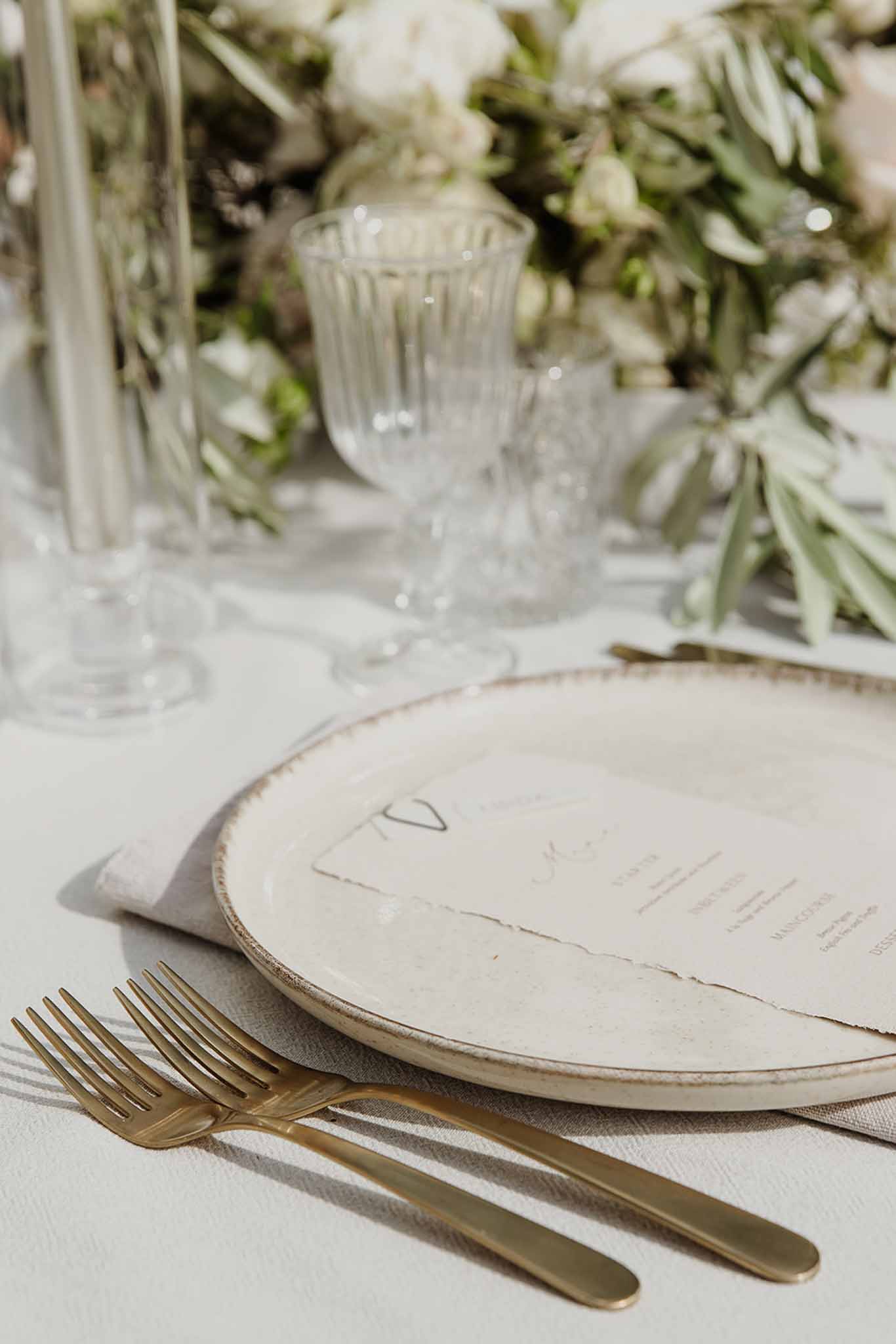 Place setting with rustic ceramic charger heart motif menu gold flatware white blooms and olive branch foliage