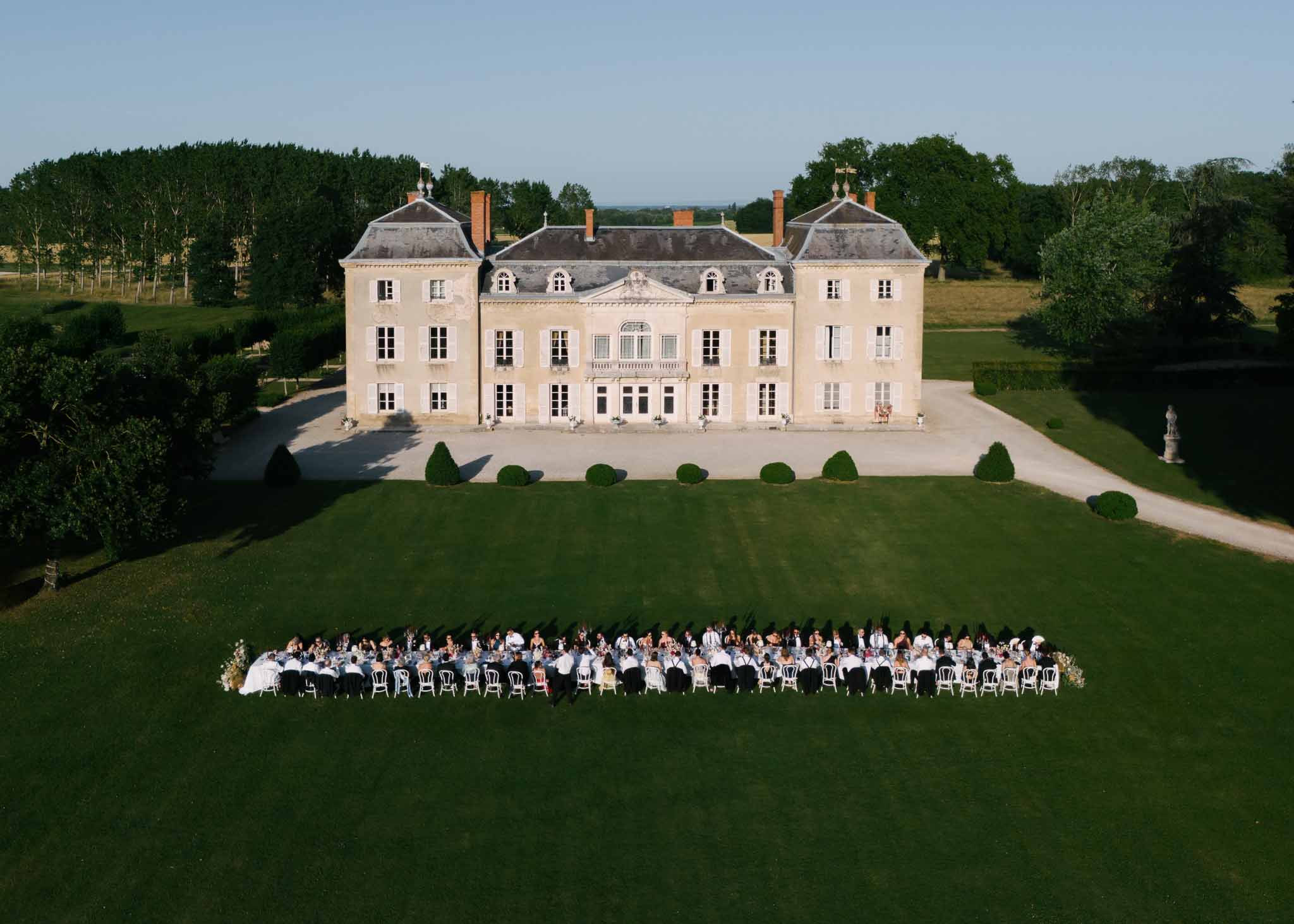 Aerial golden-hour view of 100 guests at long table on lawn before symmetrical limestone chateau