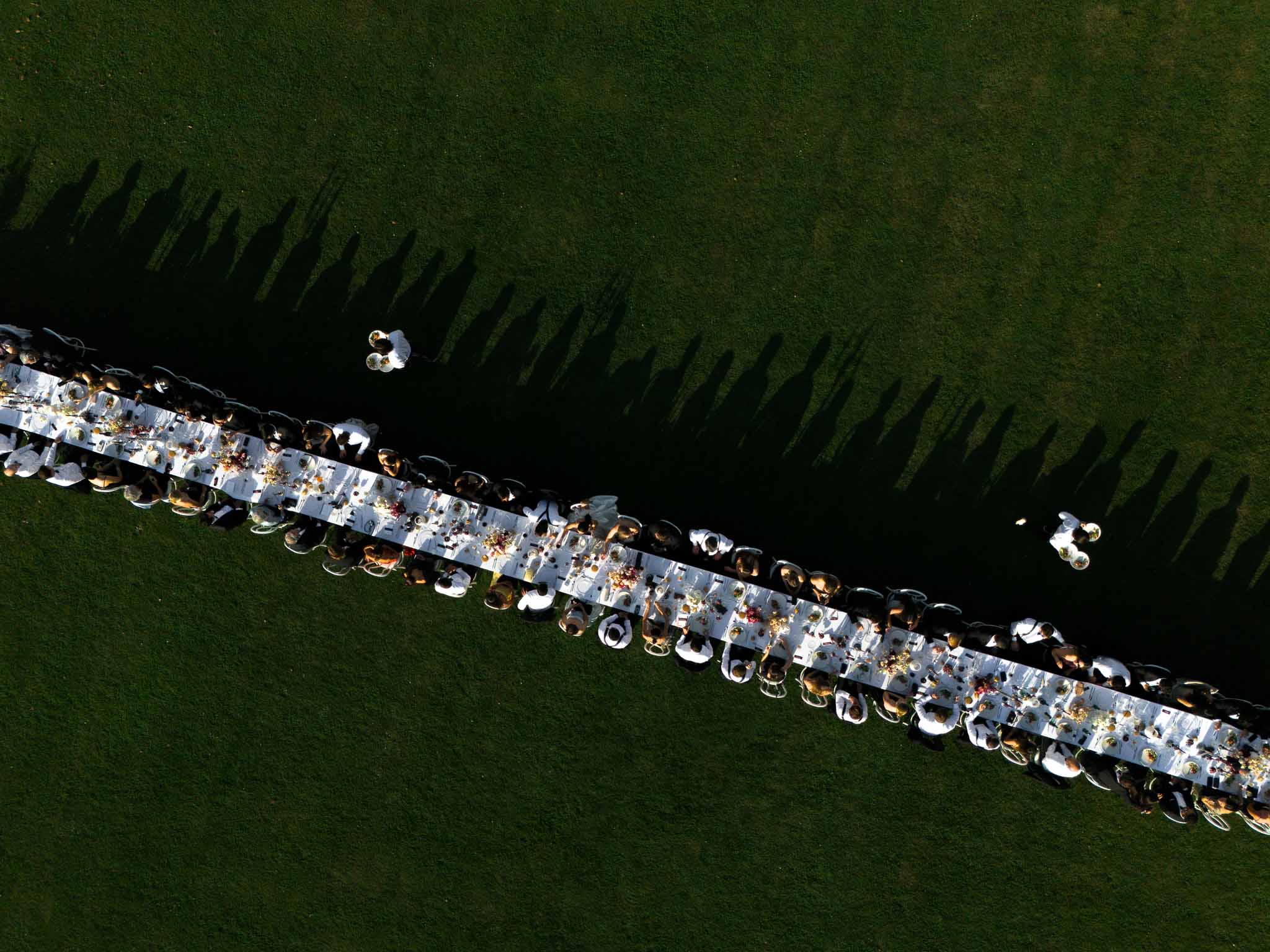 Aerial golden hour reception two long tables on lawn with sixty guests dramatic tree shadows and warm floral centerpieces