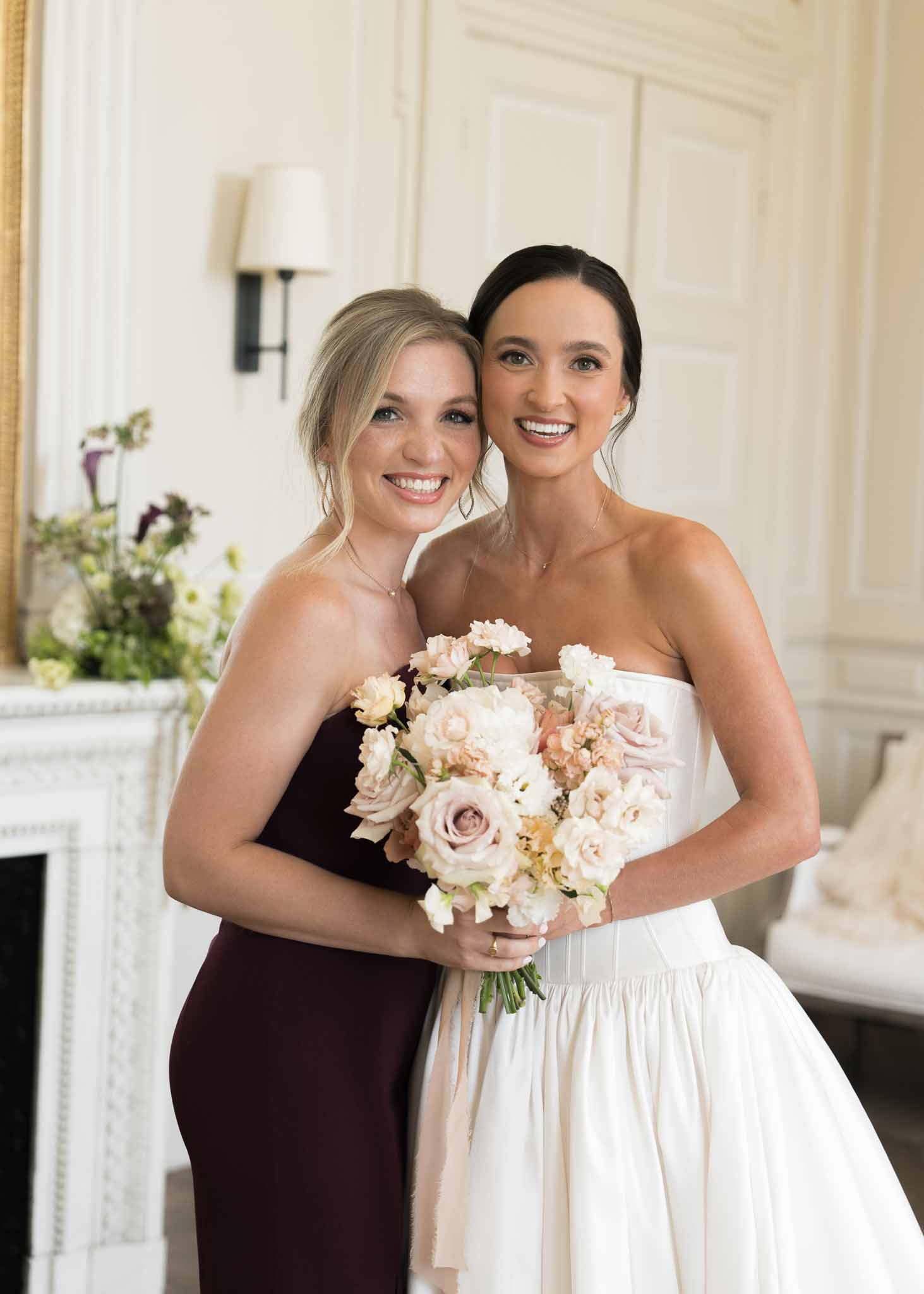 Bride with blush rose bouquet and bridesmaid in burgundy dress posing before ornate marble fireplace