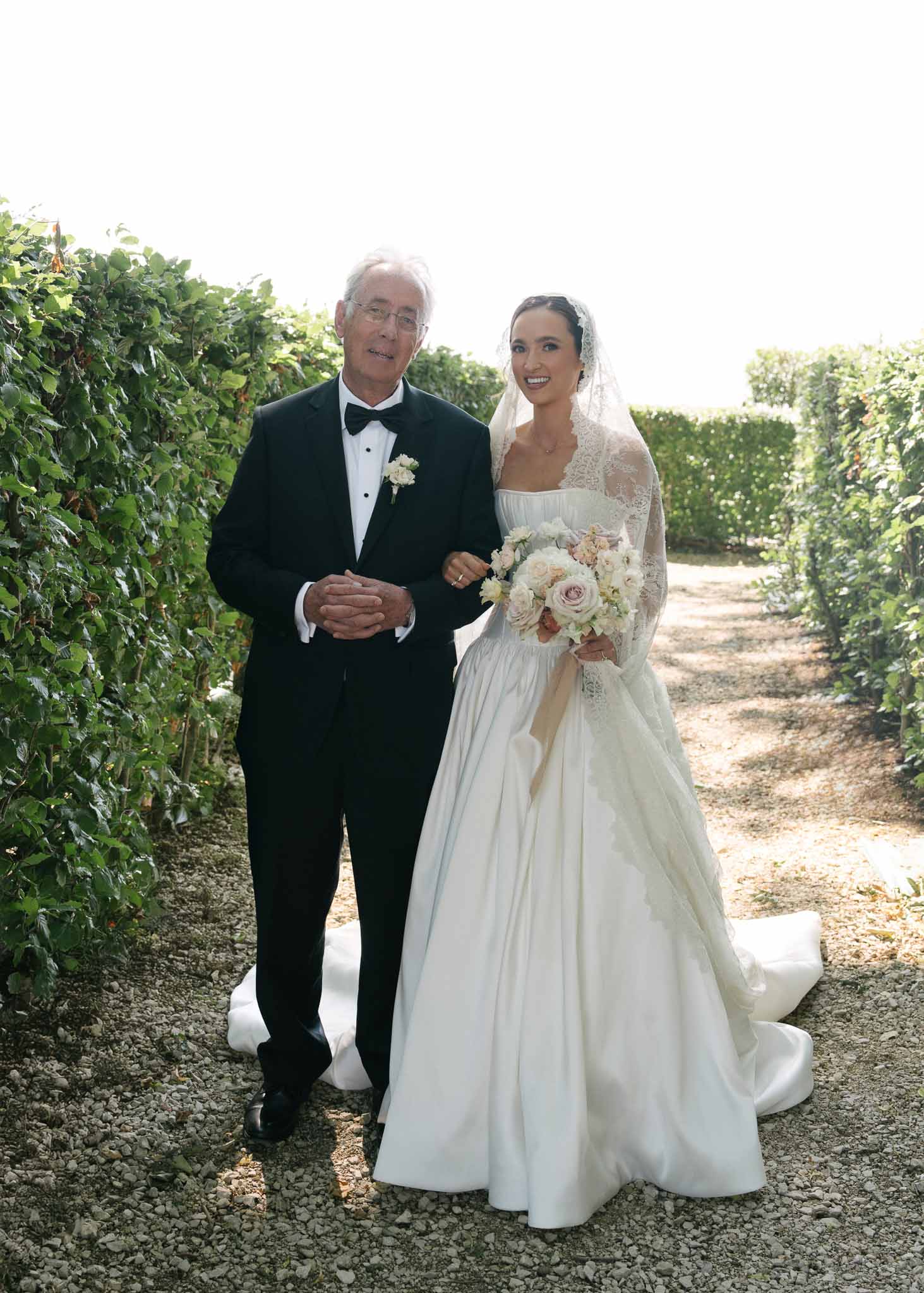 Bride in lace-sleeve ballgown walking arm-in-arm with father carrying blush and ivory bouquet
