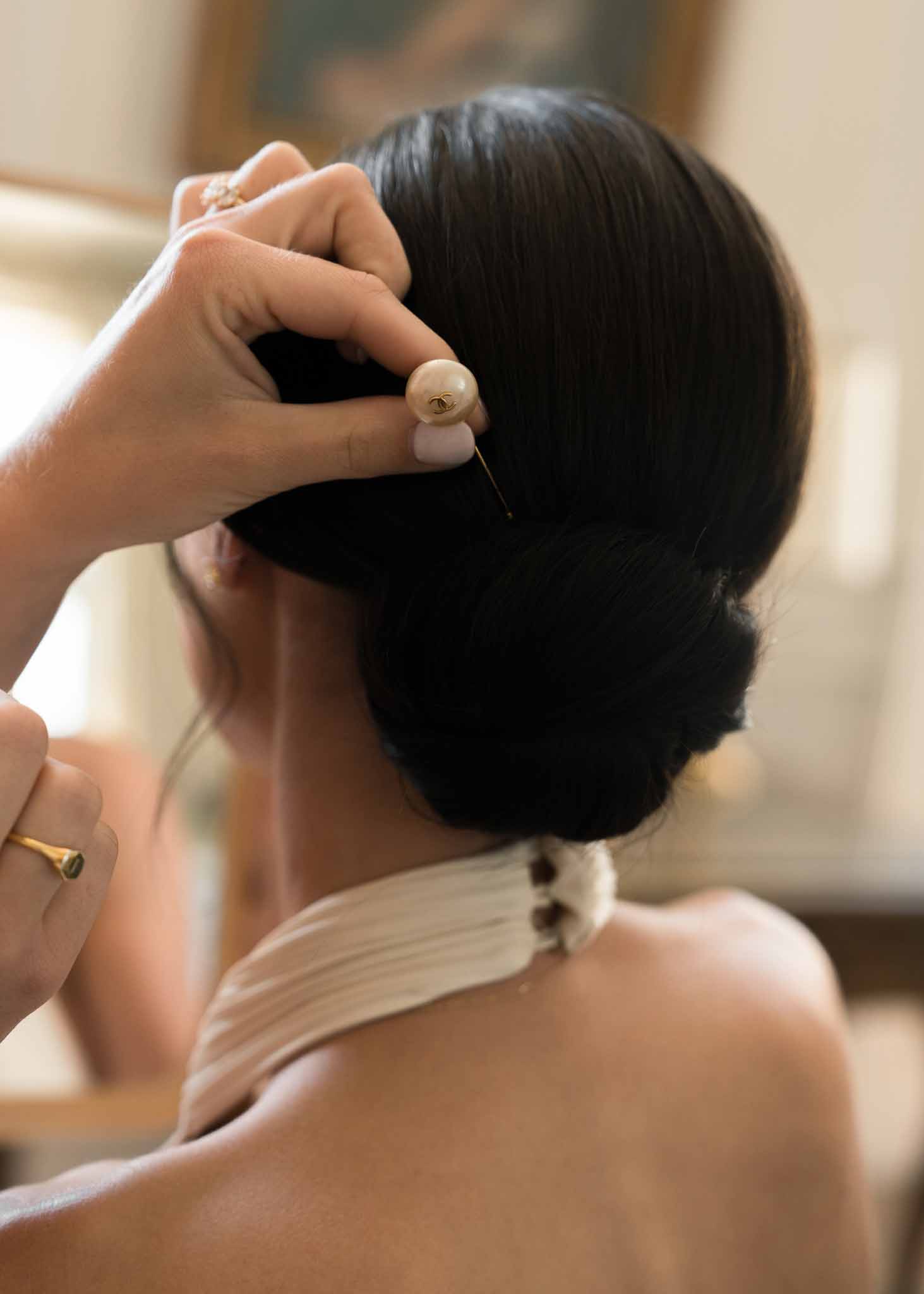 Close-up of hands pinning pearl and gold hair accessory into bride's low chignon during getting ready
