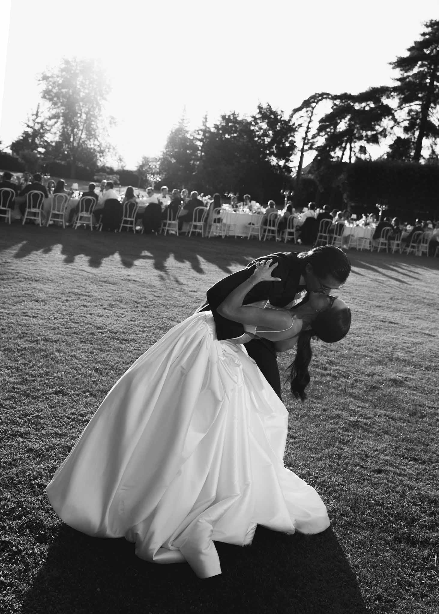 Black-and-white image of groom dipping bride for a kiss on the lawn with seated reception guests in the background