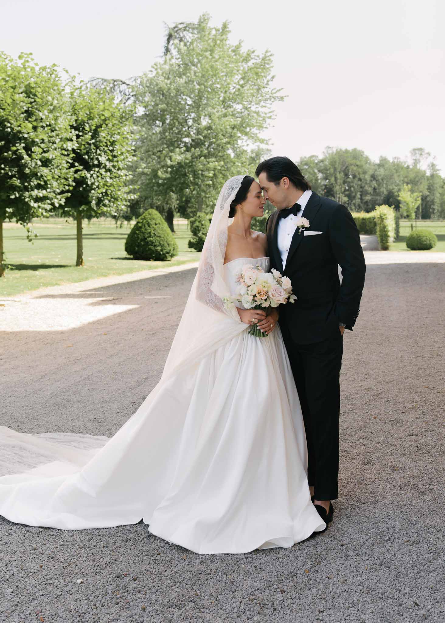 Couple forehead to forehead with blush rose bouquet and cathedral veil trailing on chateau gravel