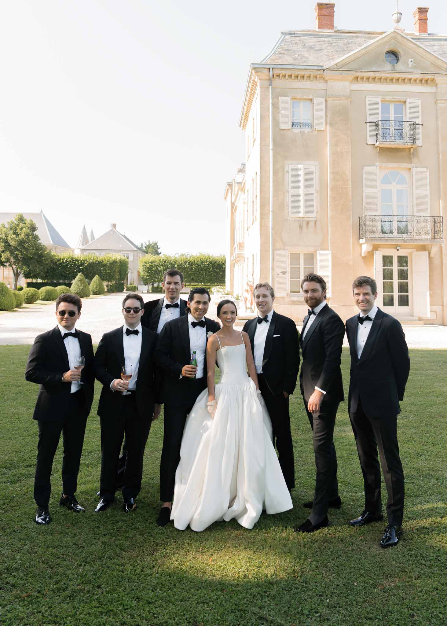 Bride in white satin ball gown posing with six groomsmen in black tuxedos on chateau lawn