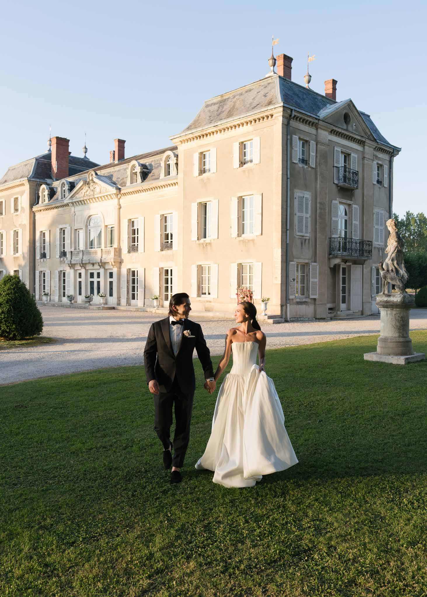 Bride in strapless ballgown and groom in charcoal tuxedo walking hand-in-hand across chateau lawn at golden hour