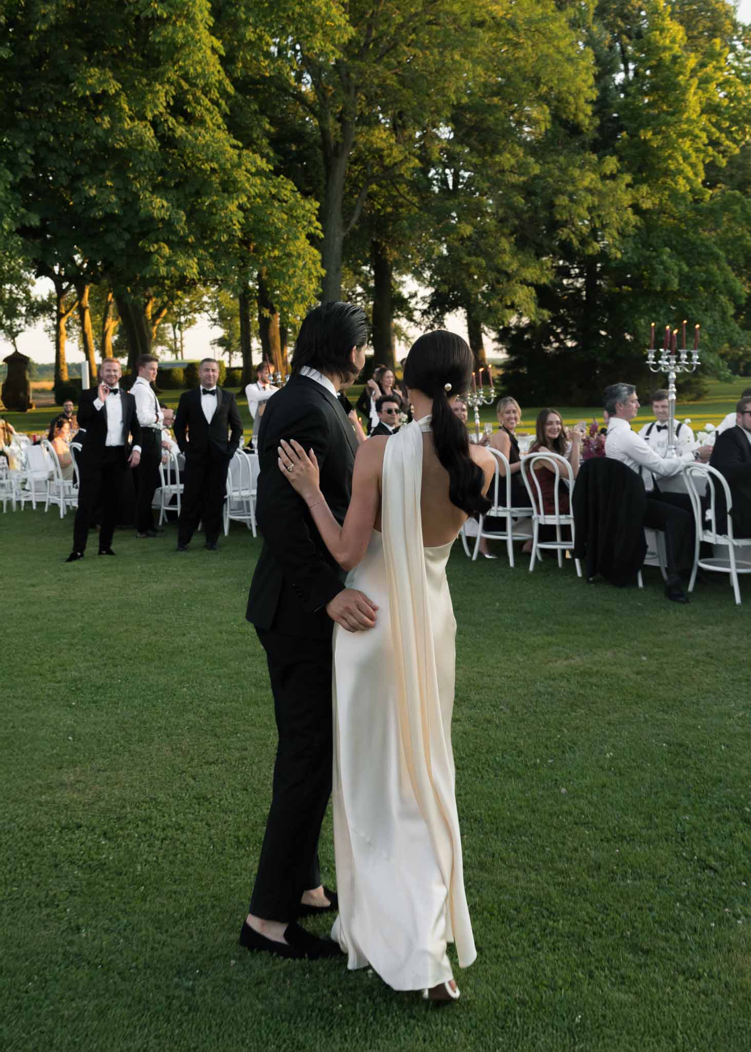 Couple from behind approaching seated guests at silver candelabra tables with red taper candles on evening lawn