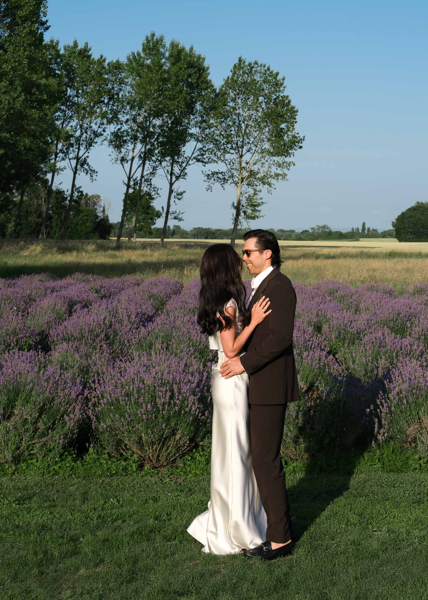 Bride and groom embracing at edge of lavender field during golden hour with wheat field beyond
