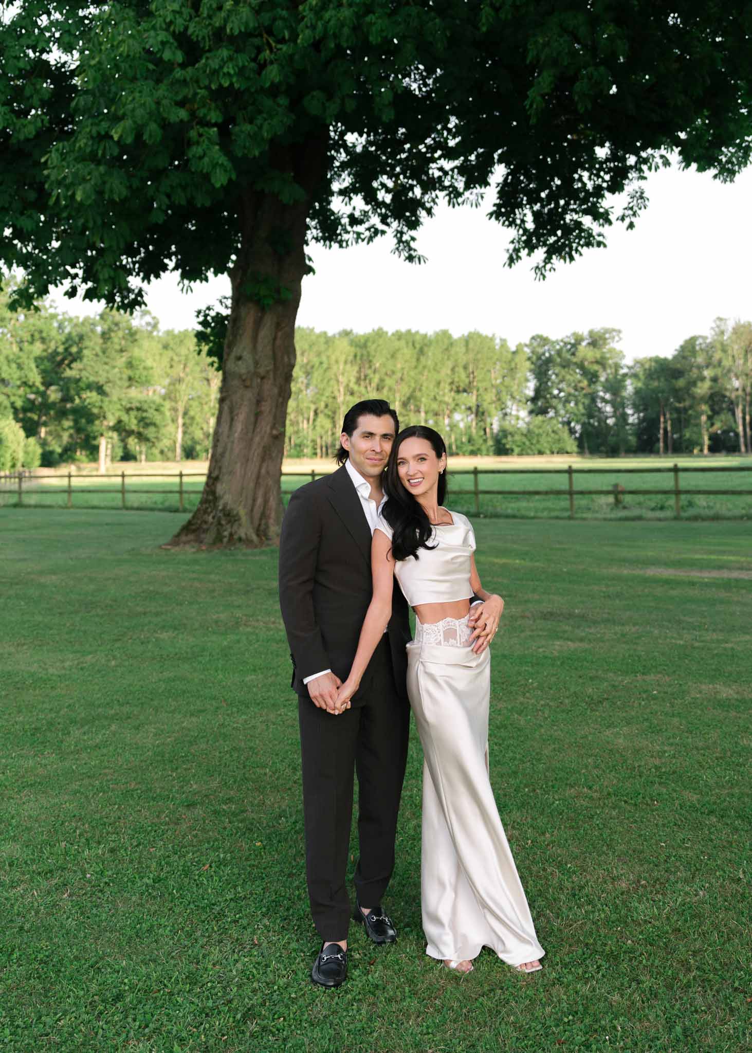 Couple holding hands under tree with bride in two-piece satin crop top and skirt at country estate