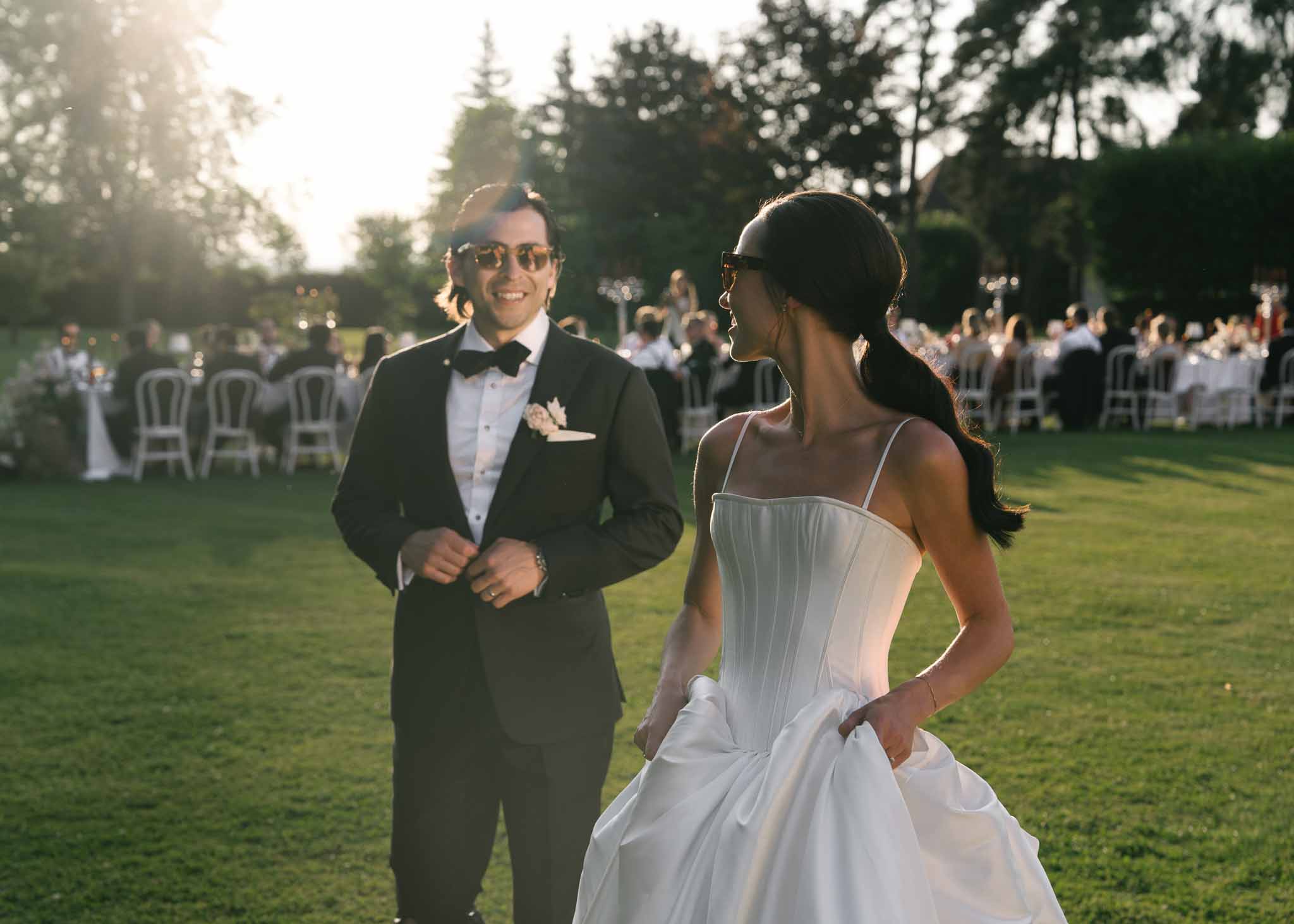 Bride in white satin ballgown and groom in black tuxedo walking across lawn during golden hour garden reception