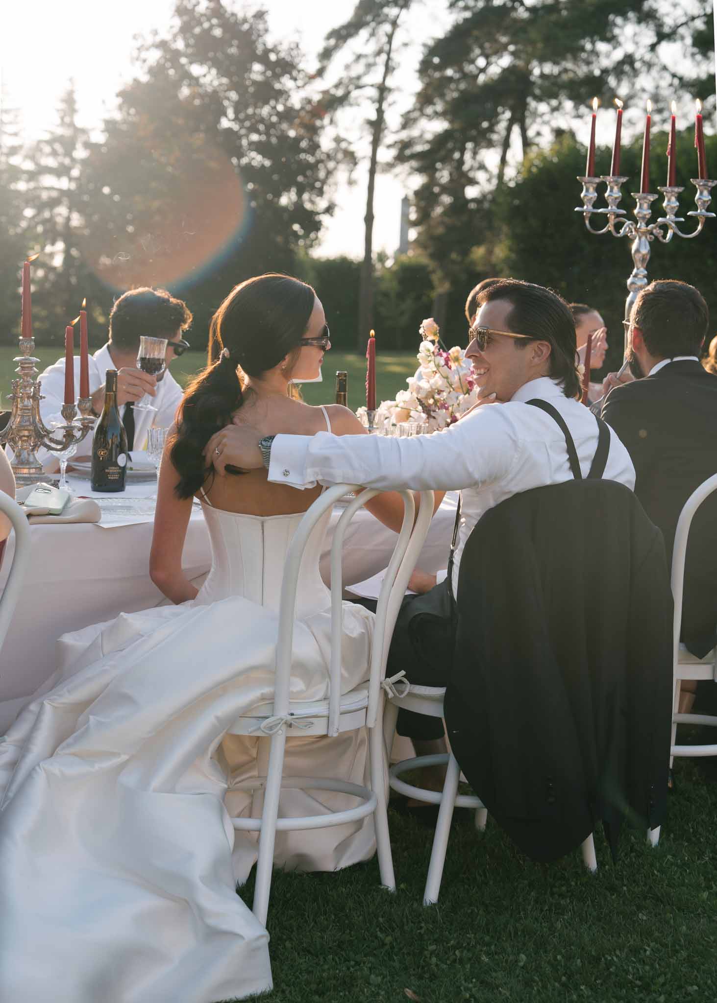 Bride and groom seated at outdoor reception table with burgundy tapers and floral centerpiece at golden hour