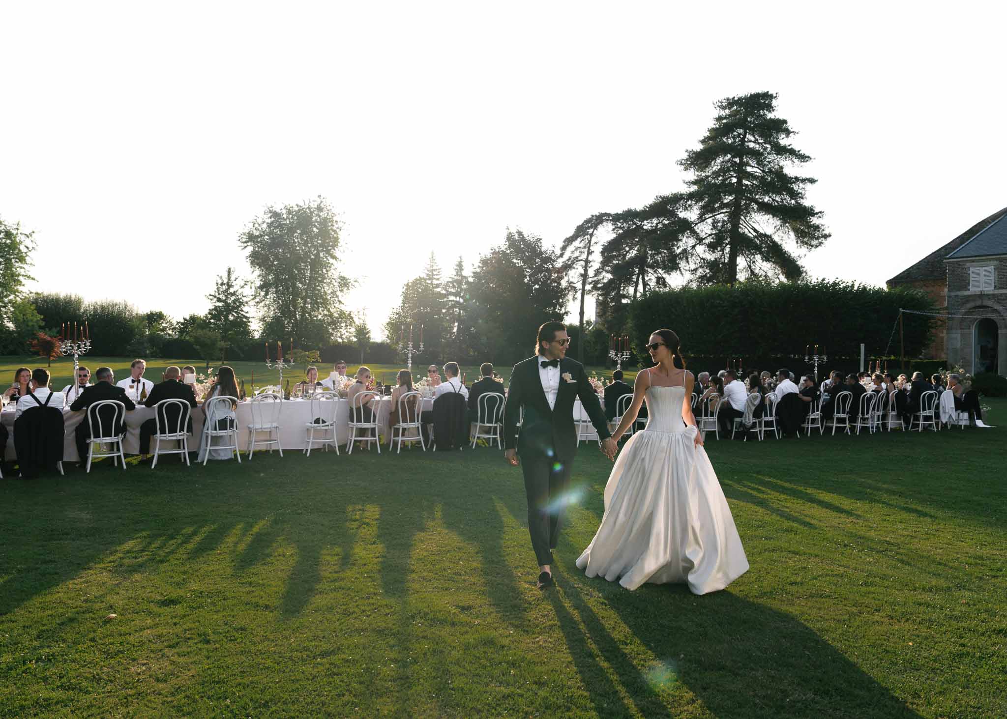 Bride and groom walk across lawn in golden hour with seated reception guests and chateau behind them