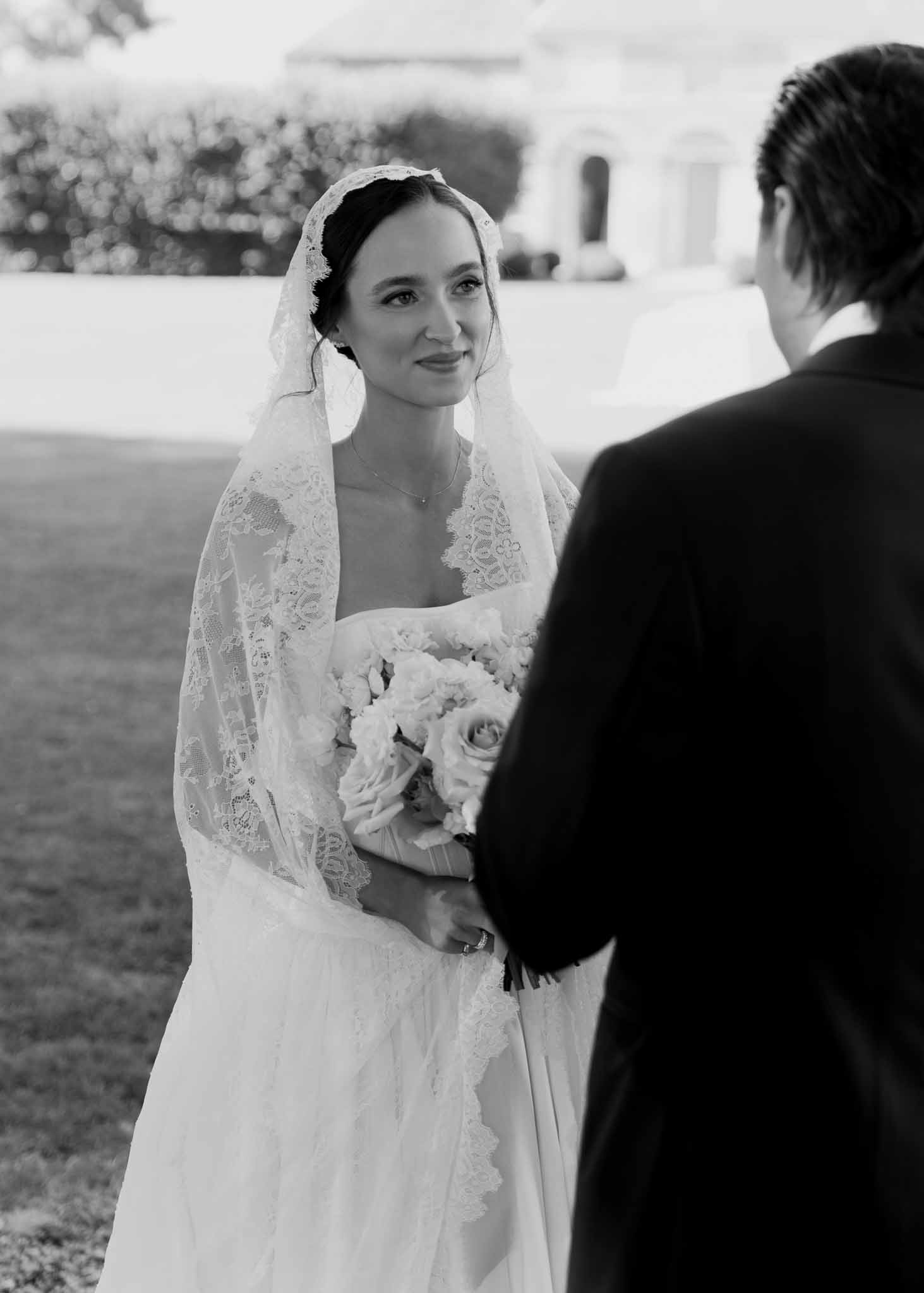 Black and white over-shoulder portrait of bride in lace gown and mantilla veil holding rose bouquet