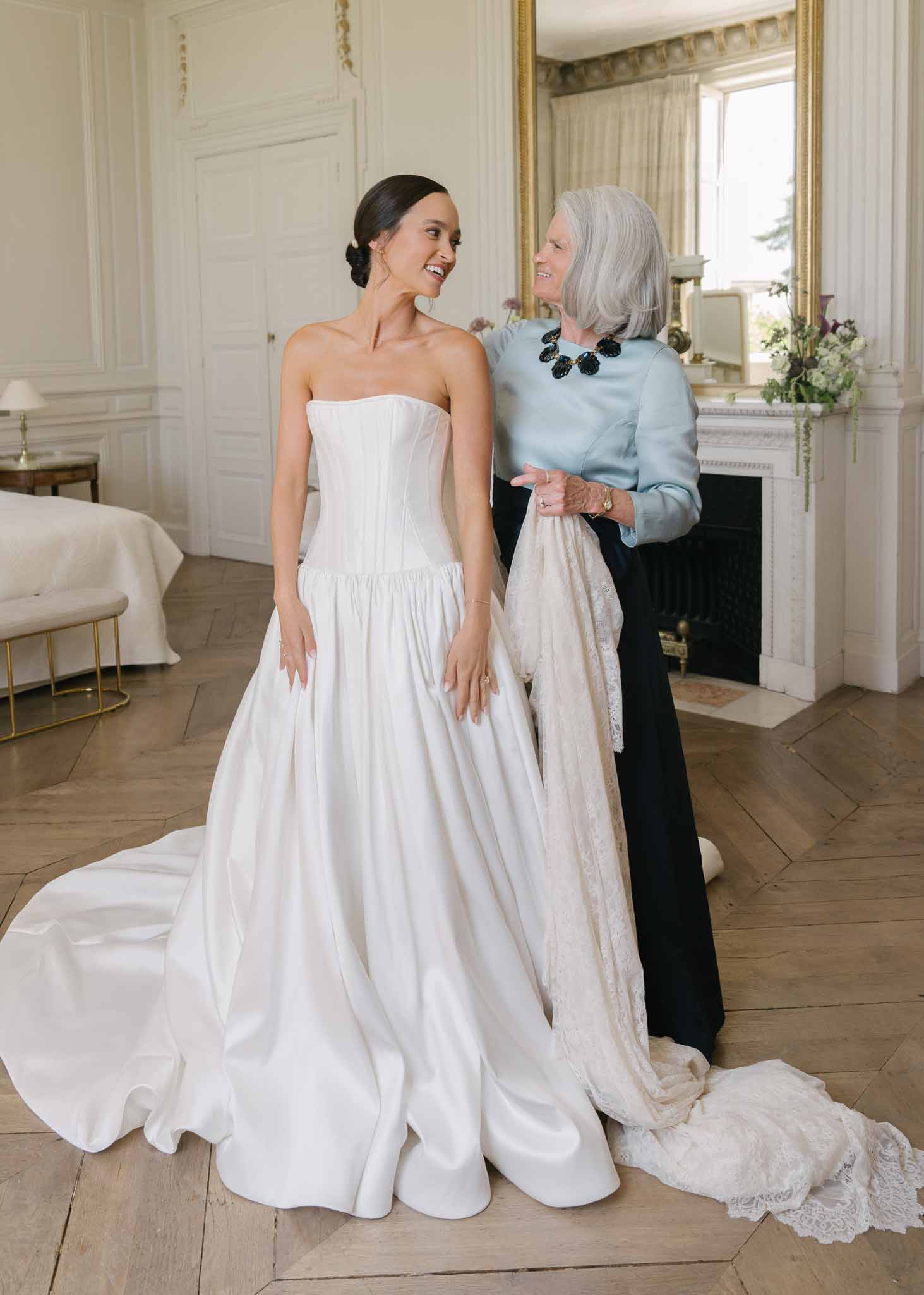 Bride and mother laughing during veil fitting in white-panelled chateau suite with gold mirror