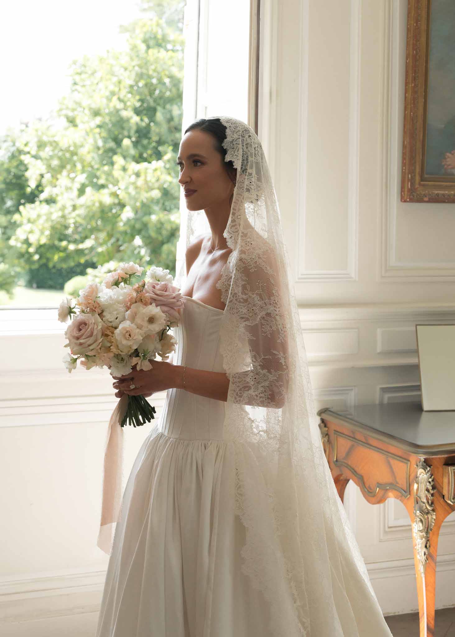 Bride in strapless ballgown with mantilla veil holds blush and white rose bouquet by window in paneled room