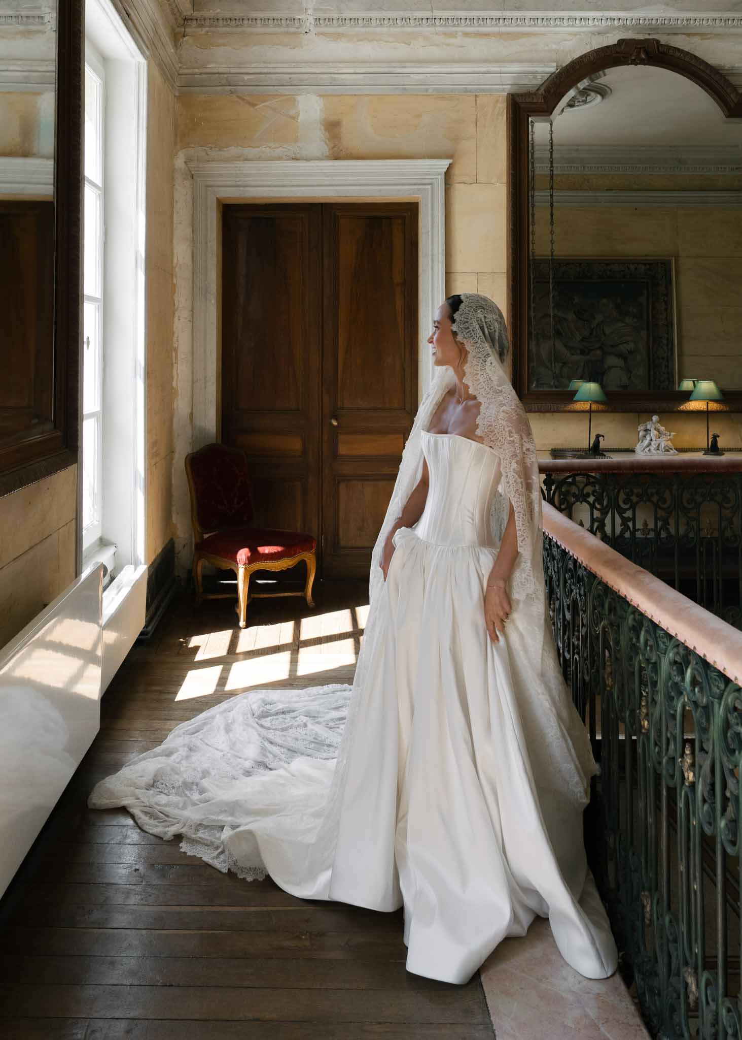 Bride in off-shoulder gown with cathedral mantilla veil on chateau gallery landing in window light