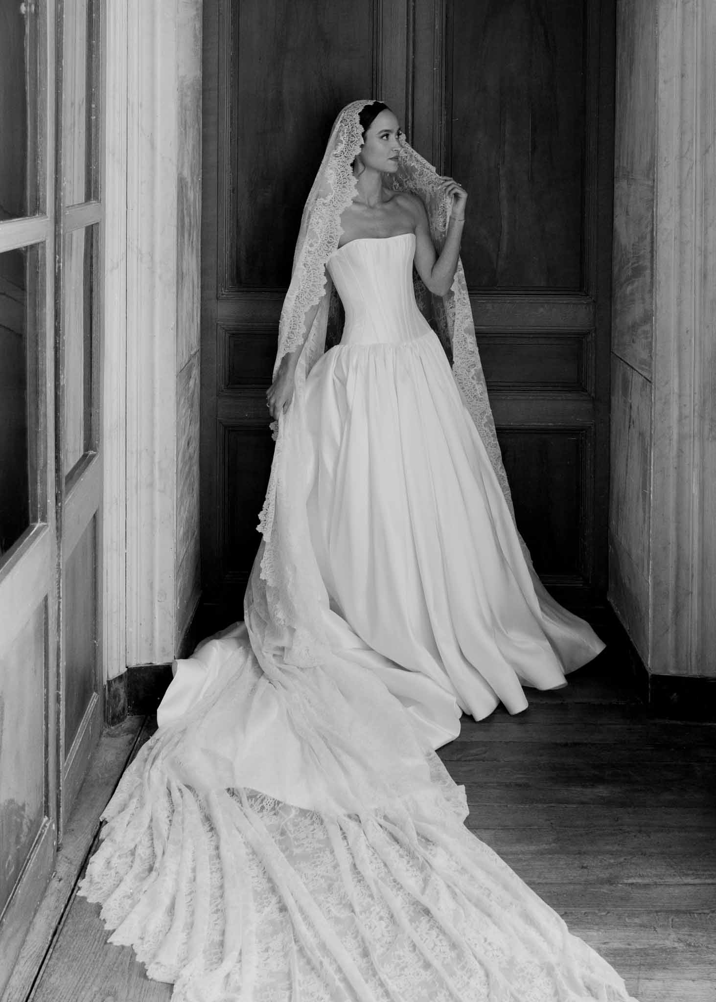 Black and white full-length of bride in strapless ballgown with lace-edged veil in dark wood doorway