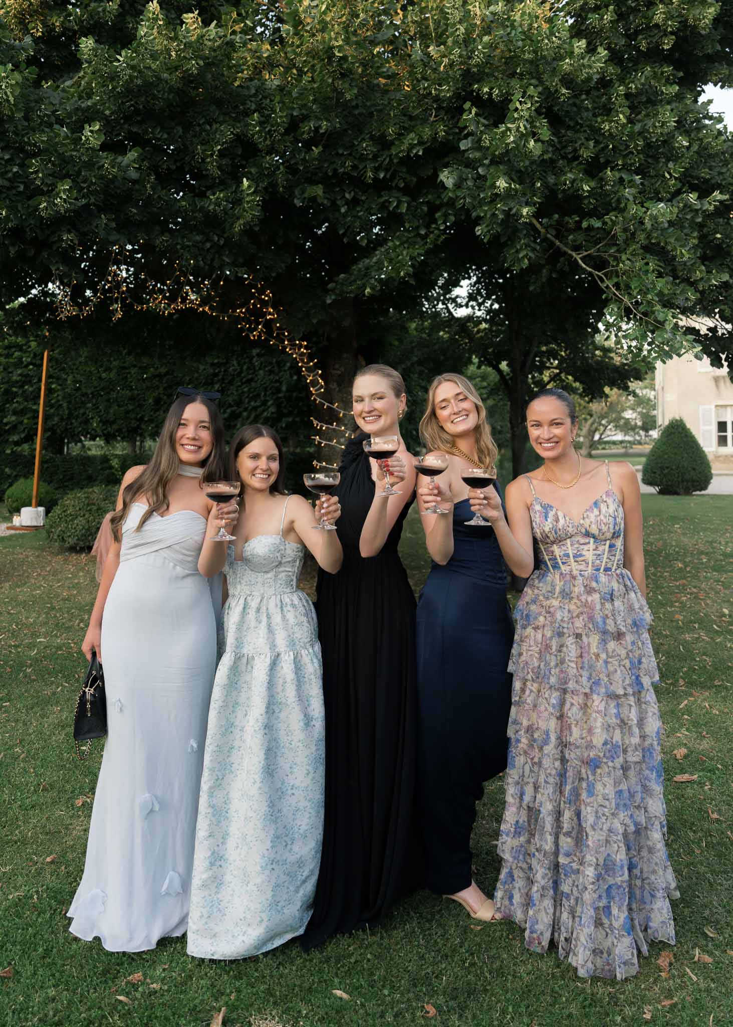 Five female guests in blue, black, and floral gowns raising red wine glasses in fairy-lit garden