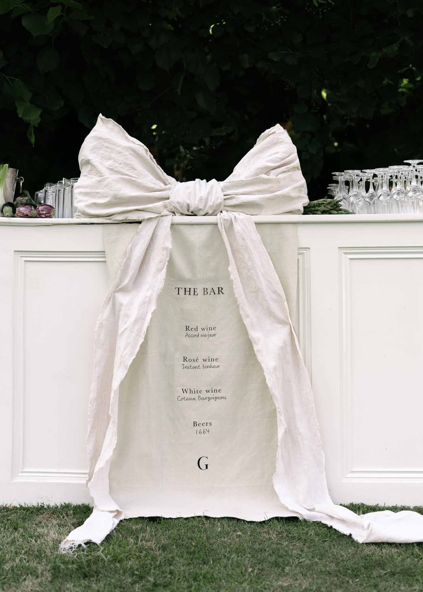 White wedding bar counter with oversized linen bow and printed drinks menu listing French wines and beer
