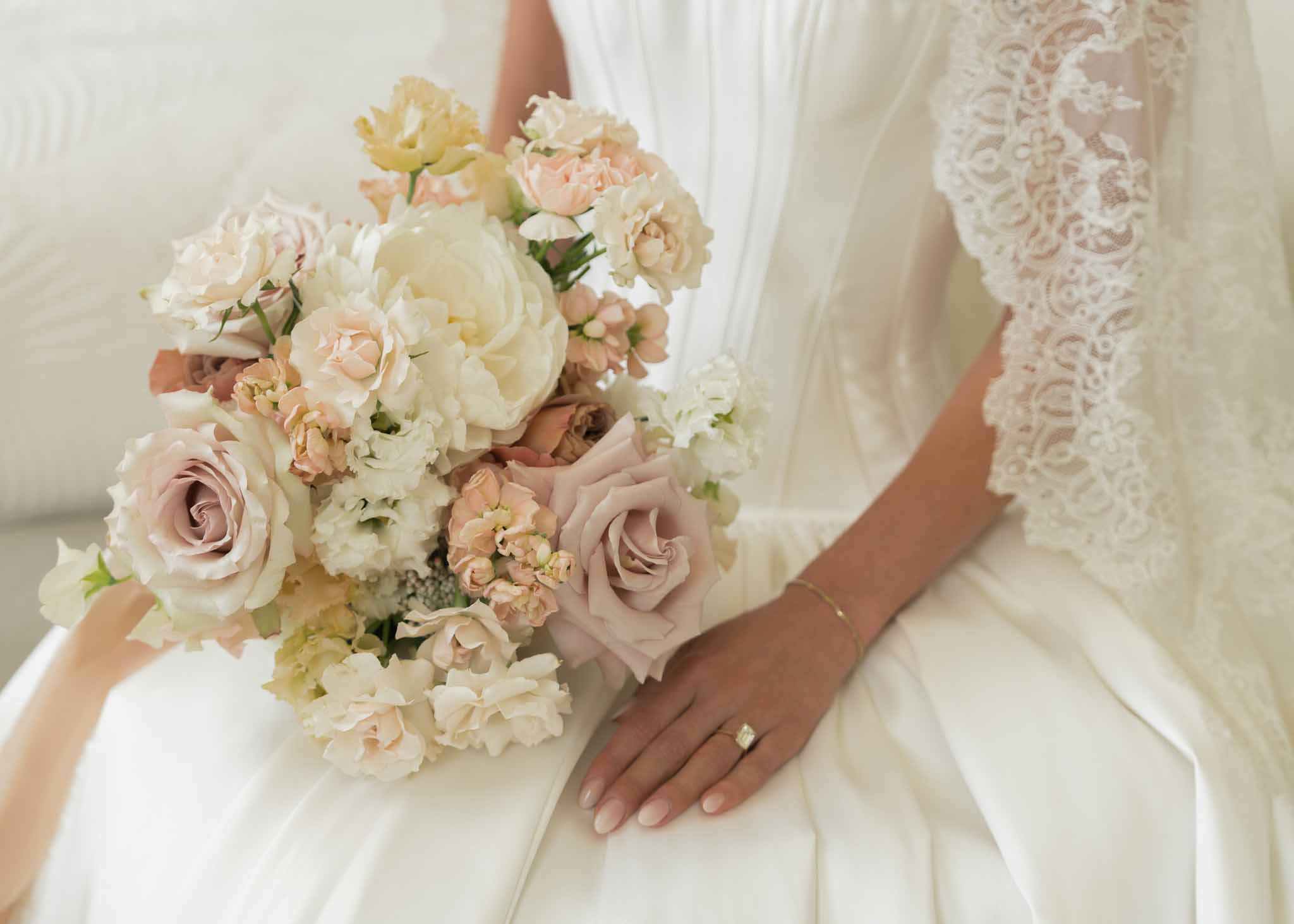 Close-up of garden-style bouquet with ivory peonies and mauve roses beside diamond ring on satin gown