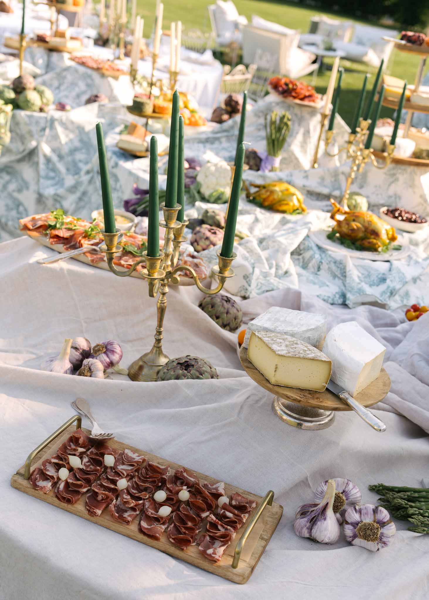 Abundant charcuterie grazing table with cheeses, artichokes, and gold candelabra with green tapers