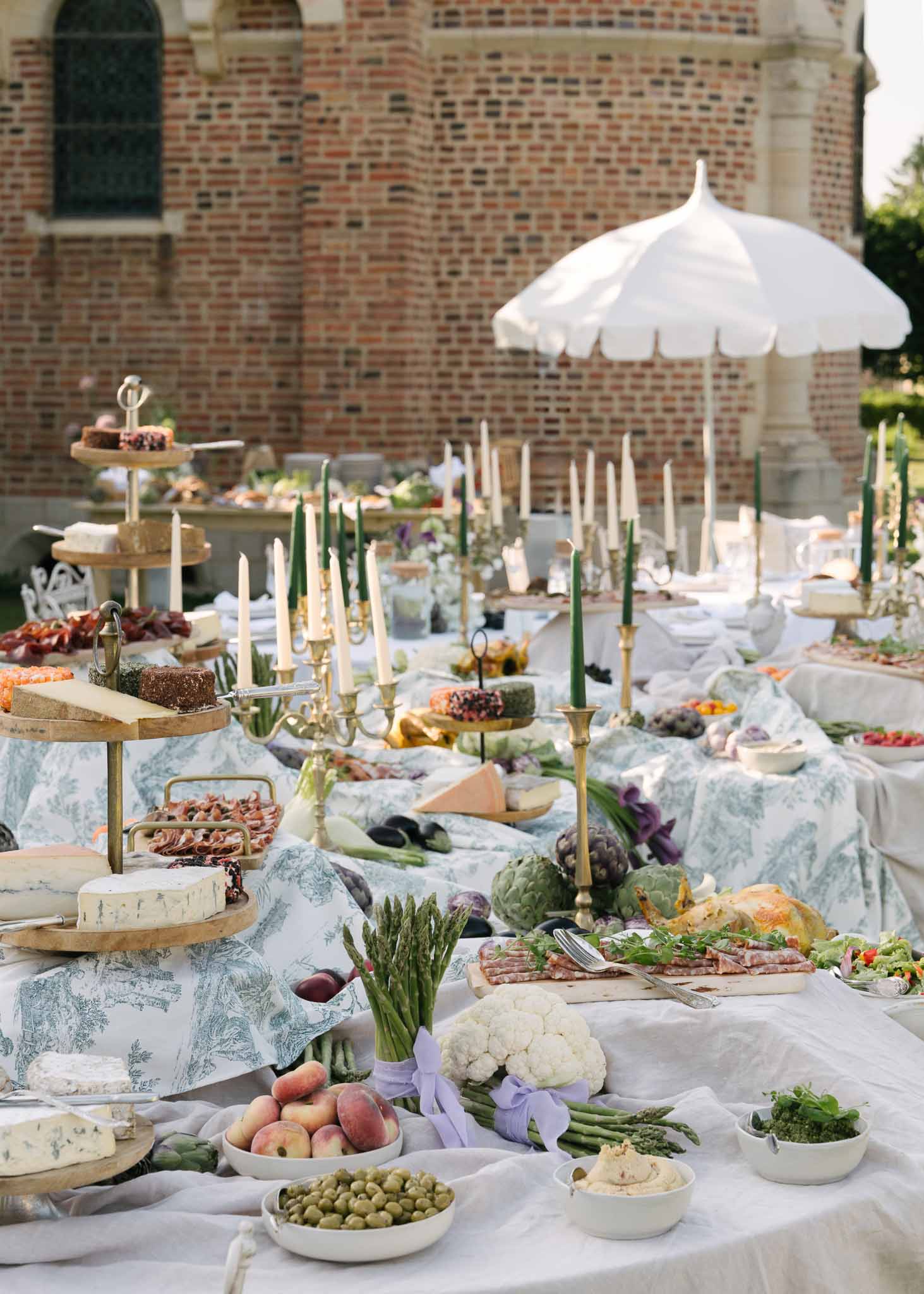 Grazing table with toile overlay, cheeses, charcuterie, and brass candelabras before red brick chateau
