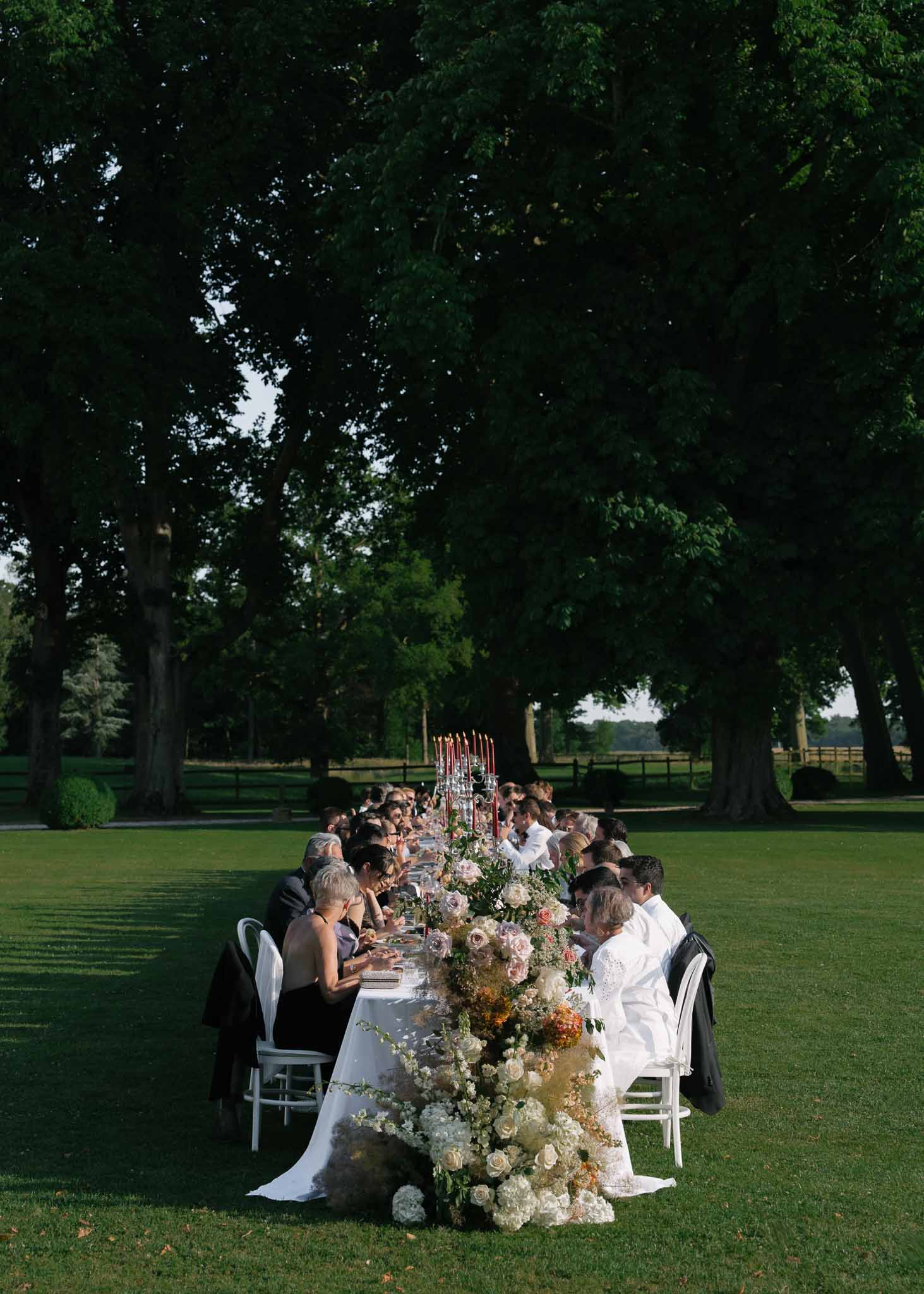 Long table with cascading cream and dusty rose floral runner and burgundy tapers beneath mature trees