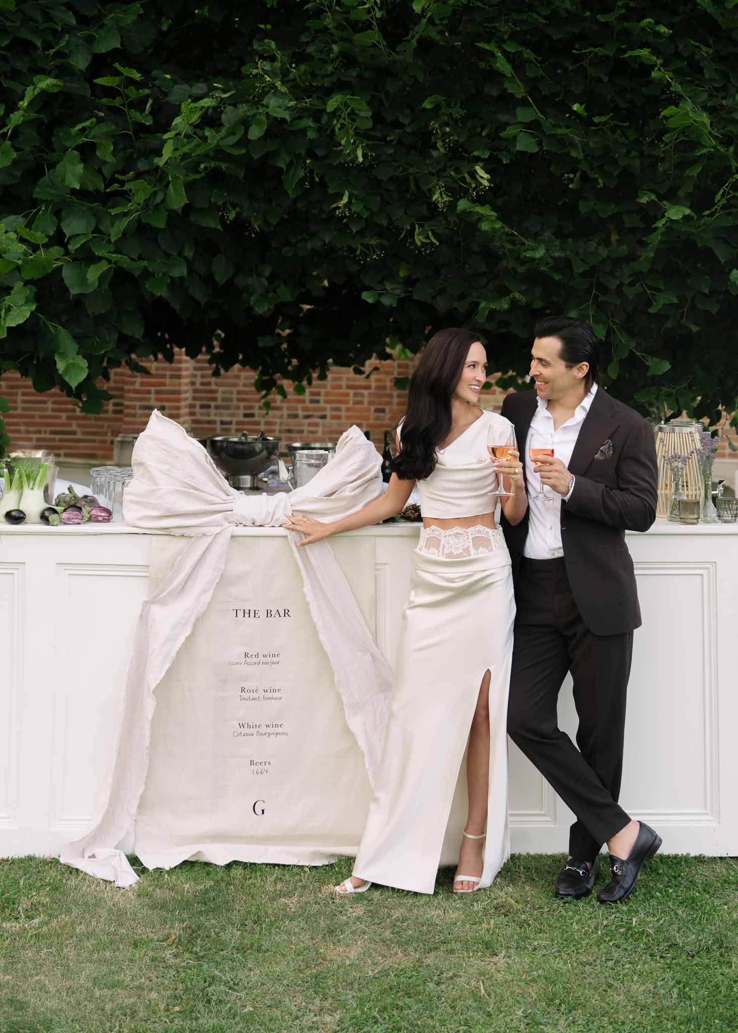 Bride in two-piece ivory outfit and groom at white bar station during outdoor cocktail hour
