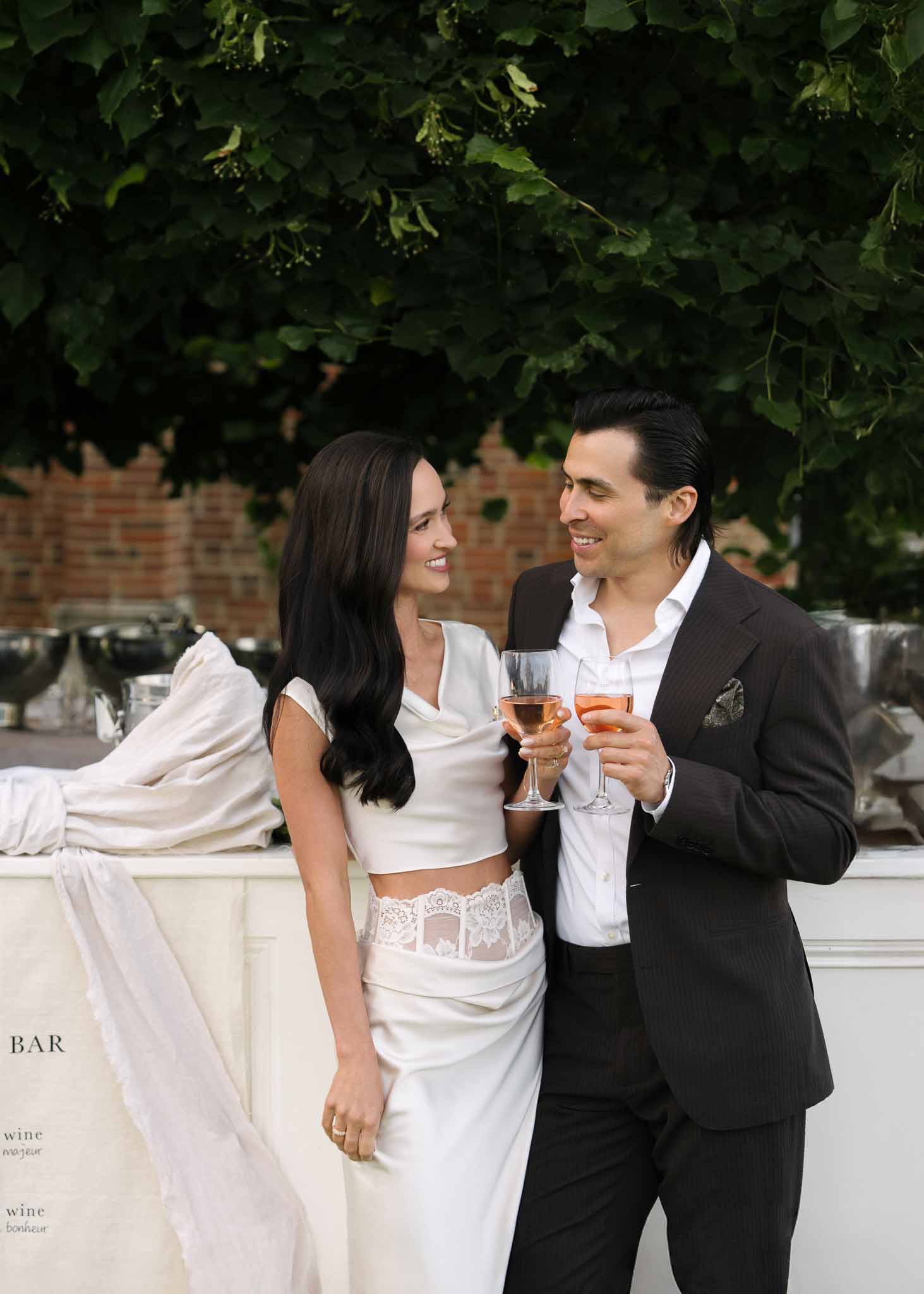 Couple holding rose wine glasses at white outdoor bar station with Venue Exterior brick backdrop