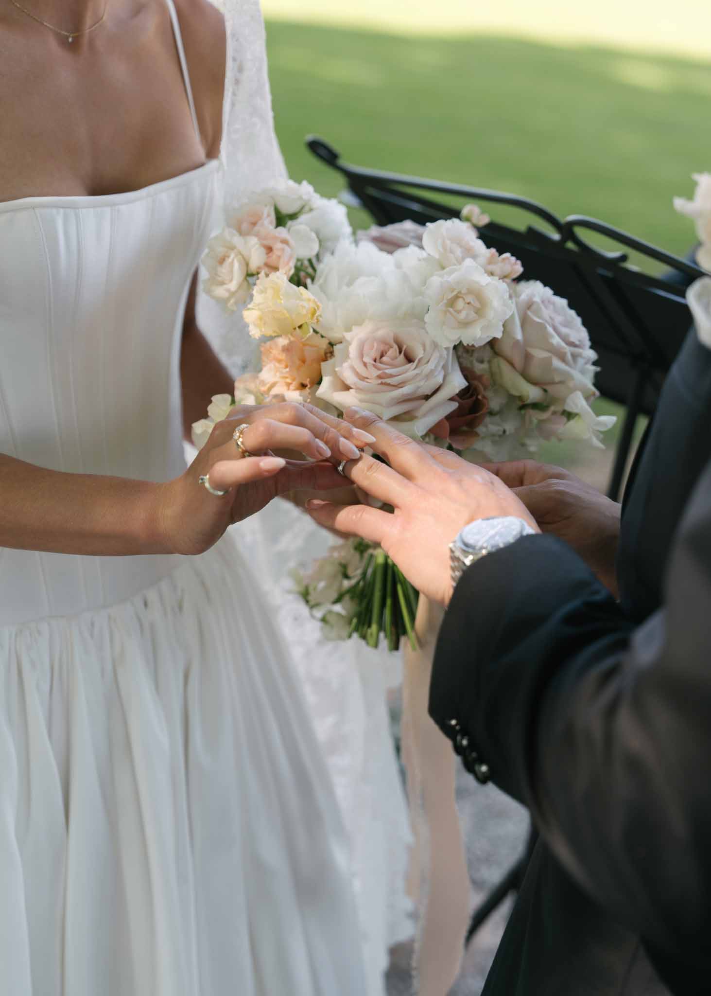 Close-up ring exchange with bride holding ivory peony and blush rose bouquet at outdoor ceremony