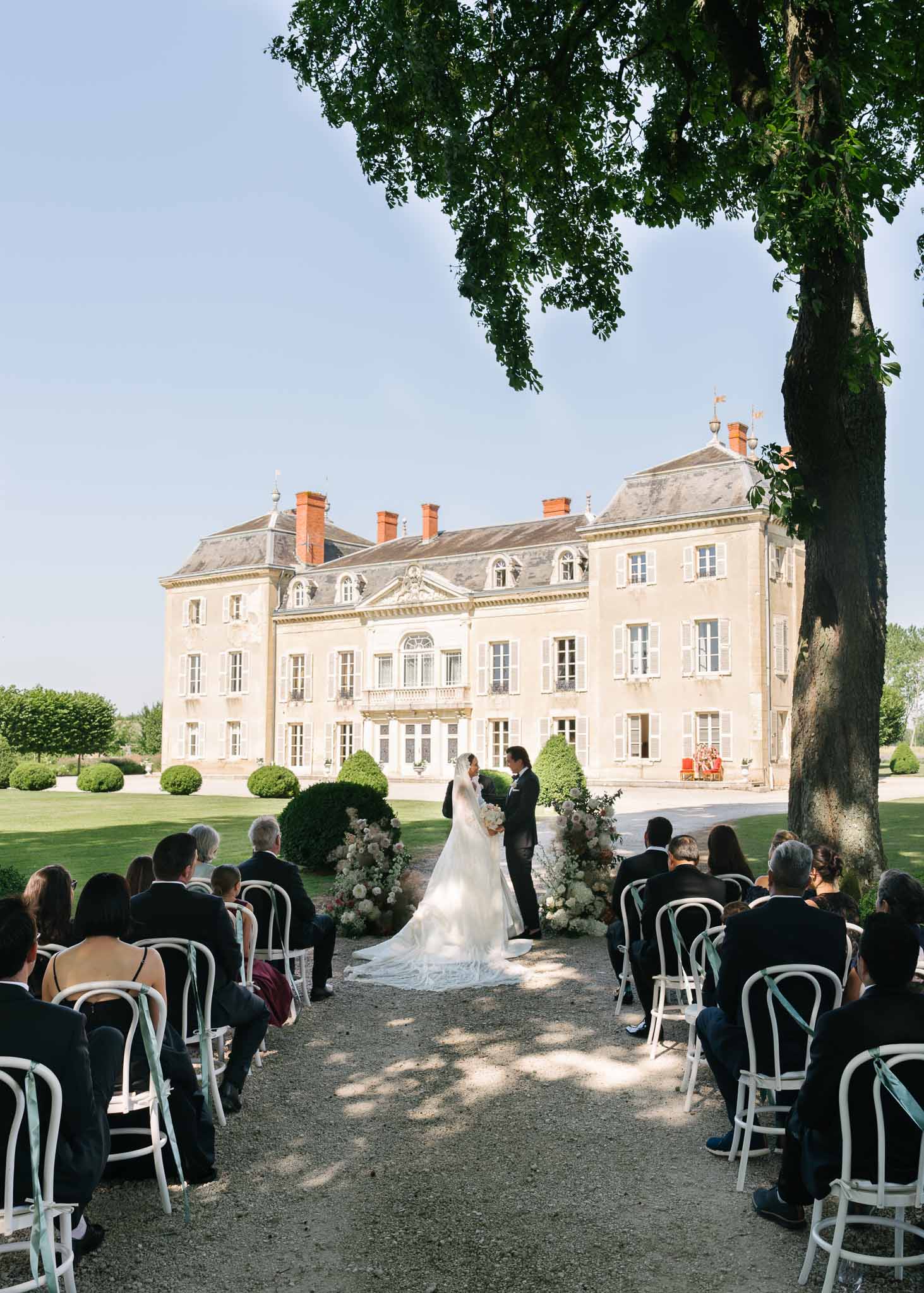 50 guests at chateau ceremony with white floral arch and sage ribbon-tied bentwood chairs