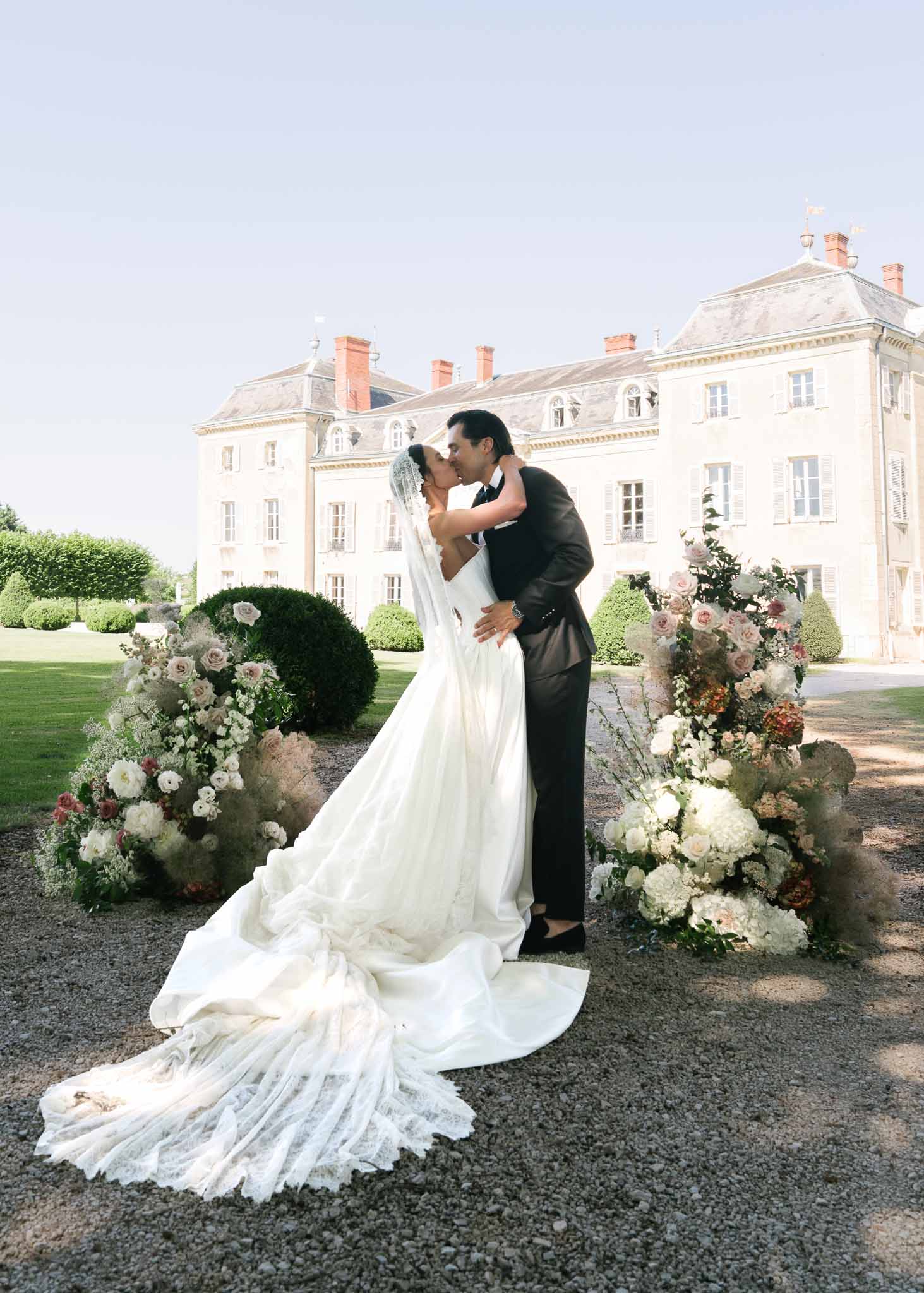 Bride and groom kiss on chateau forecourt flanked by ground-level floral arrangements with dahlias and pampas grass